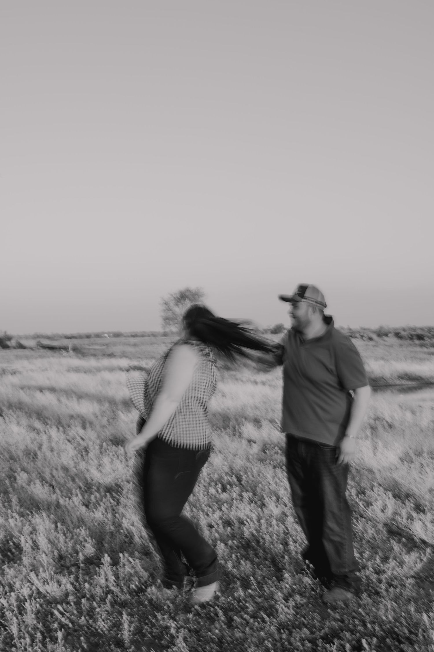 Black and white photo of a couple dancing and laughing together in an open field, womans hair blowing in the wind.