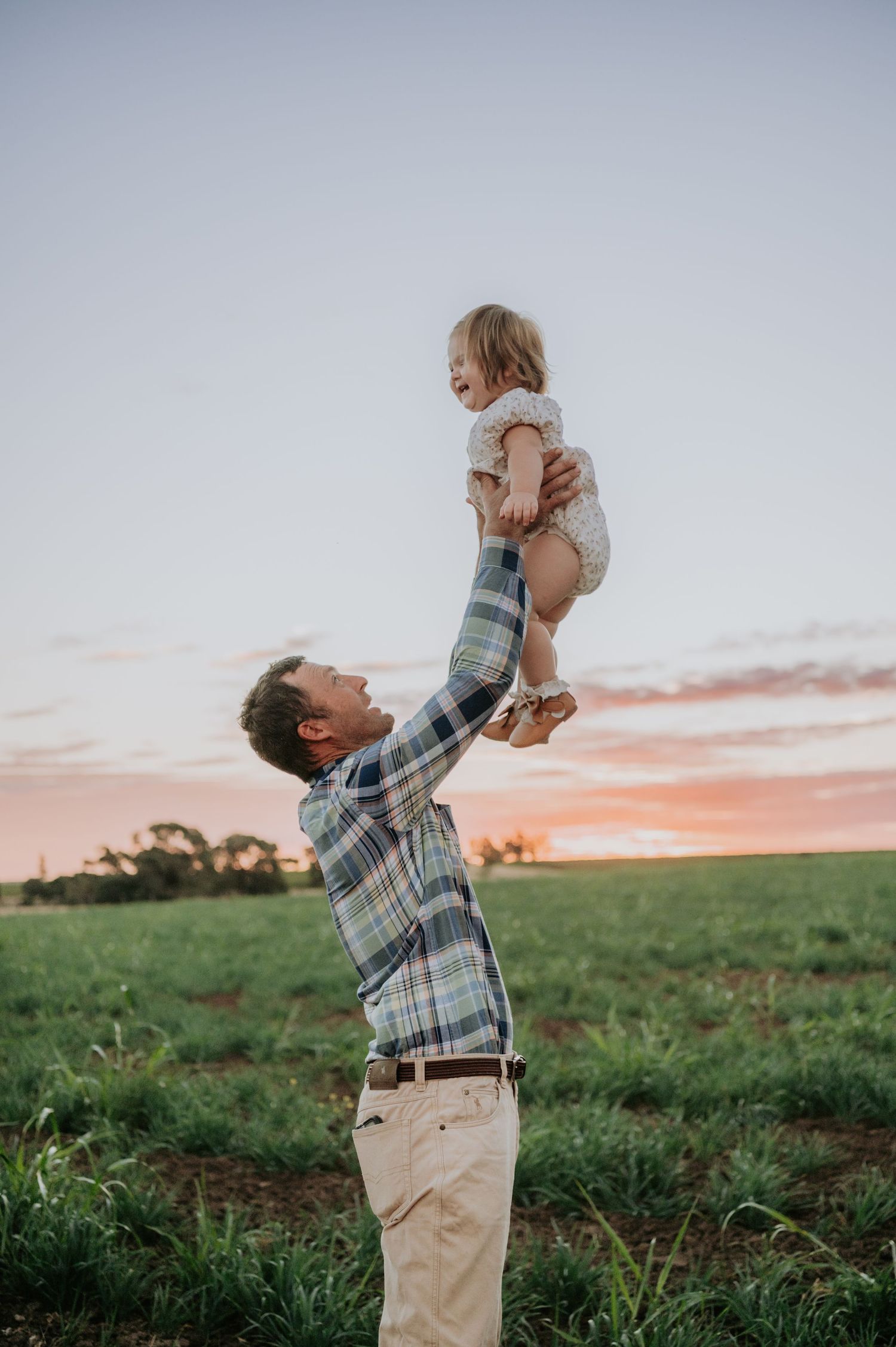 Father lifting laughing toddler high in the air at sunset in a green field.