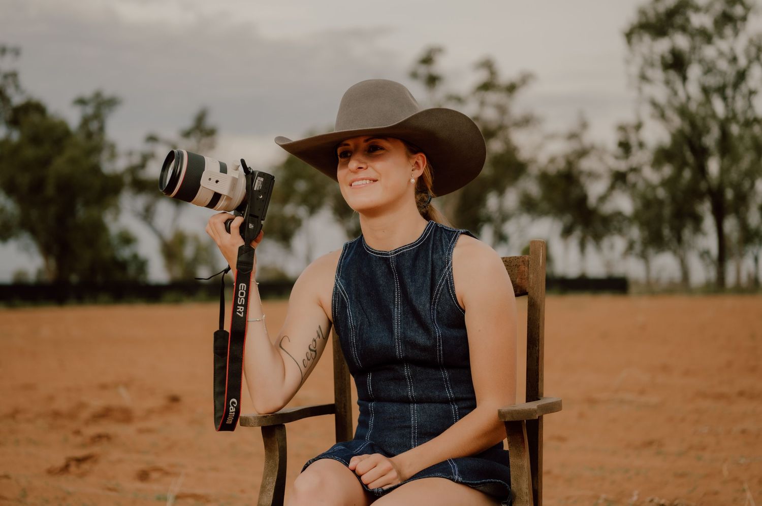 A photographer wearing a cowboy hat and dark vest takes photos in a dusty outback setting during golden hour.