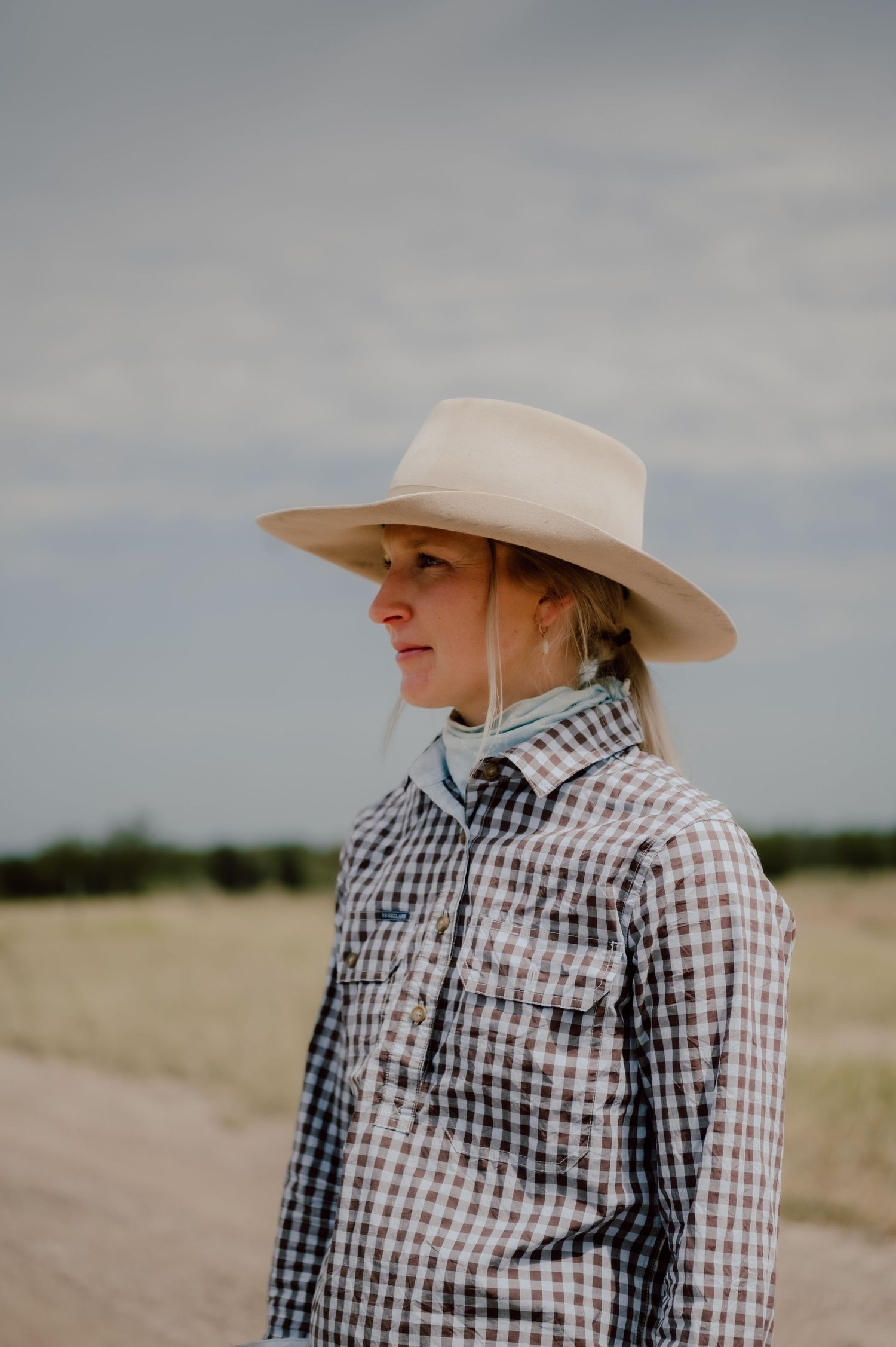 Person wearing gingham shirt and cowboy hat in rural setting.