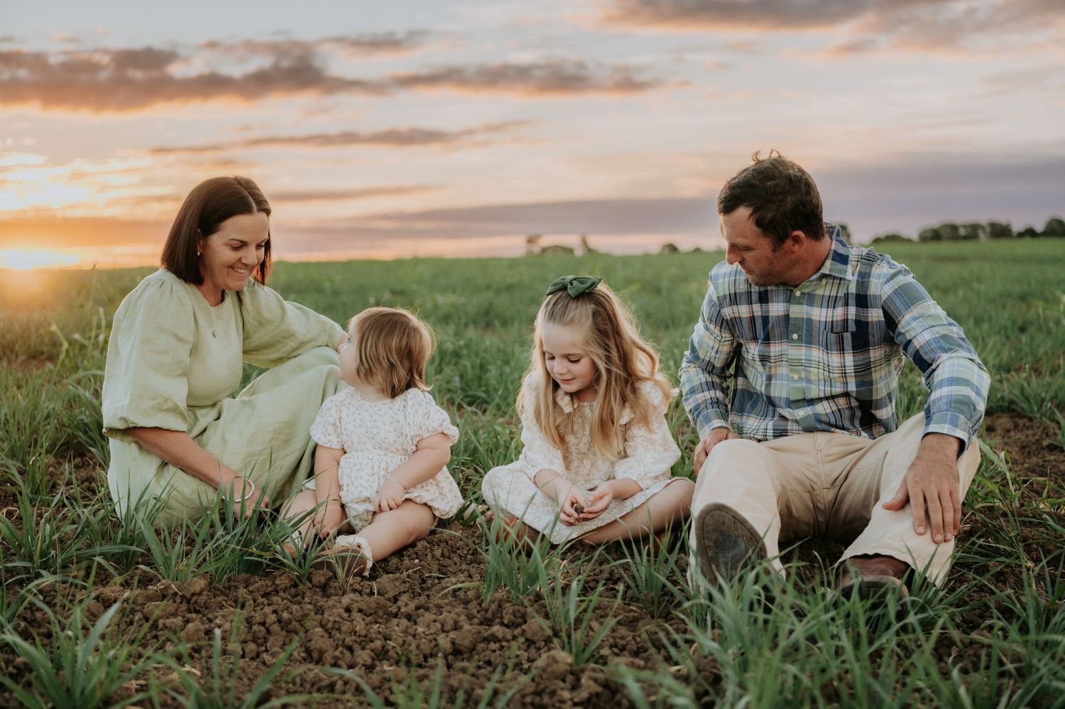 Family of four sitting in a green field at sunset, parents and two young girls smiling together outdoors.