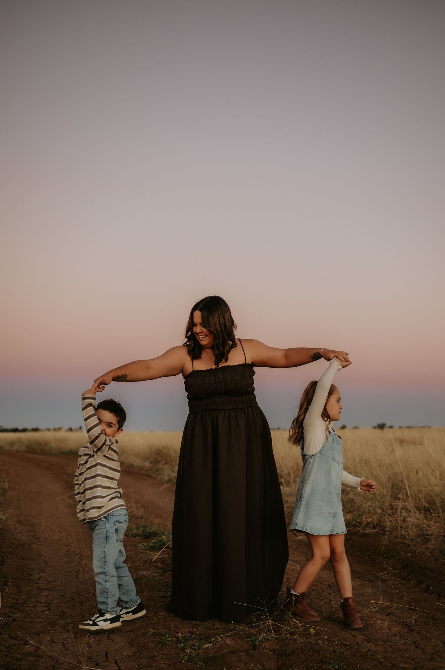 A mother in a black dress holds hands with two children dancing in a field at sunset.