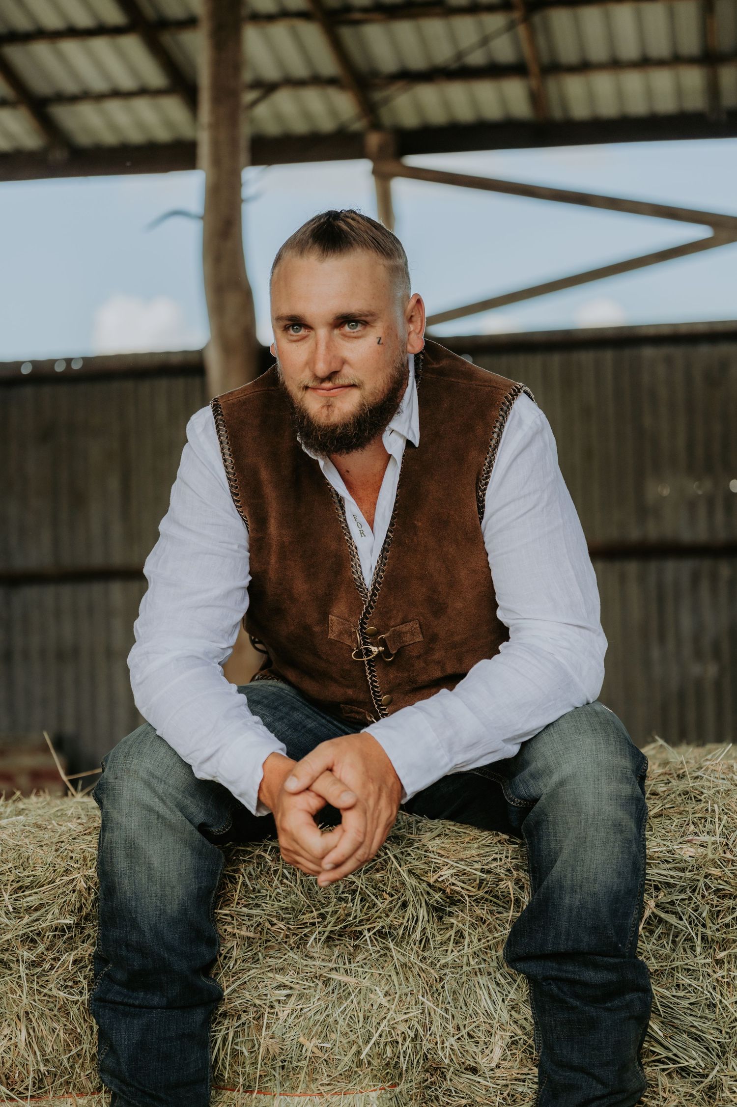 Man in brown leather vest and white shirt sits on hay bale in rustic barn setting.