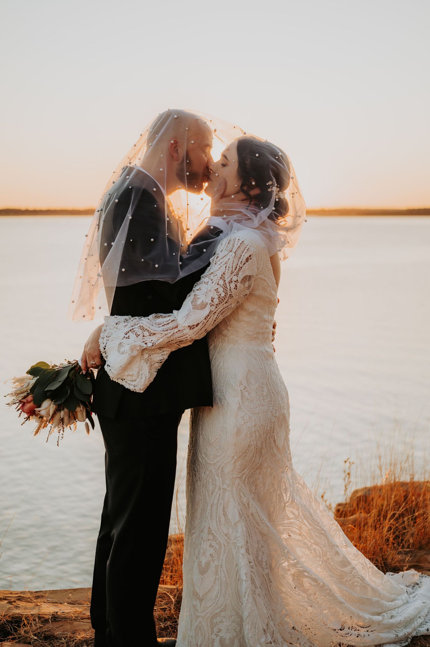 Bride and groom kissing under a veil at sunset by a lake, bride holding a bouquet in a lace wedding dress.