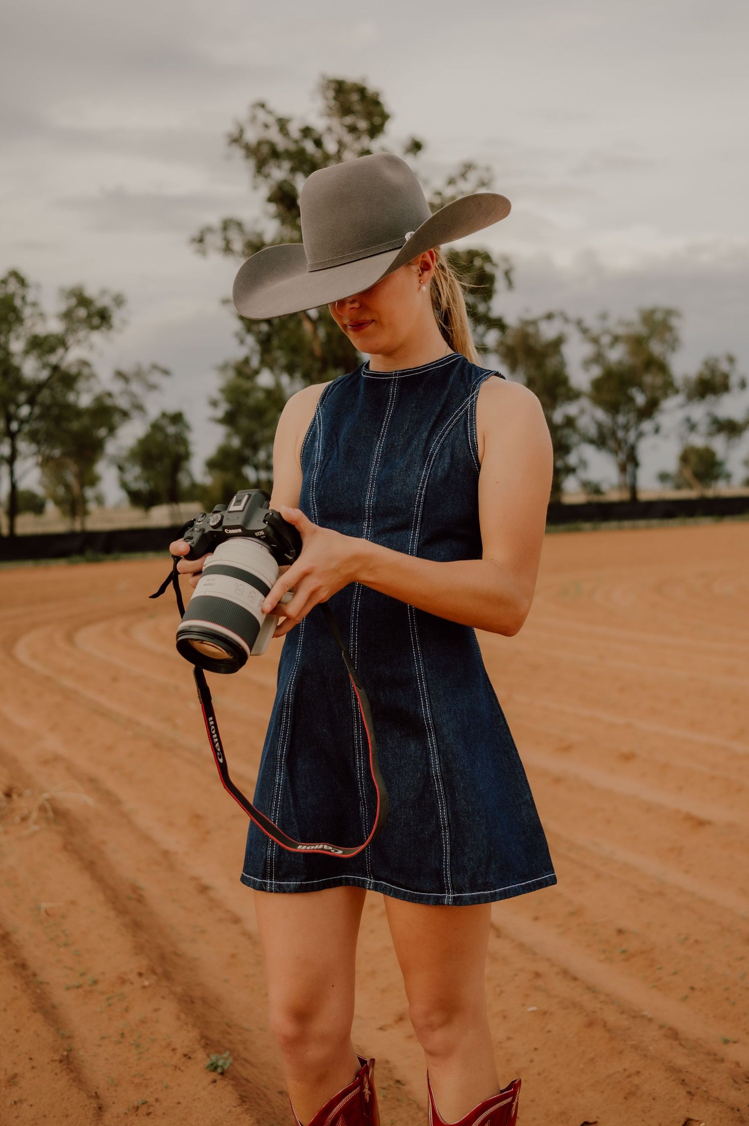 A child in a denim dress and wide-brimmed hat explores red dirt farmland while holding a camera.