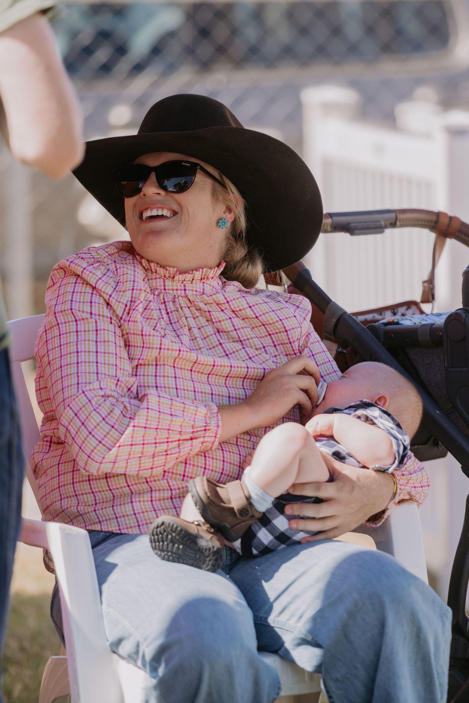 Smiling woman in black cowboy hat and pink plaid shirt sits in white chair holding a baby at an outdoor event.