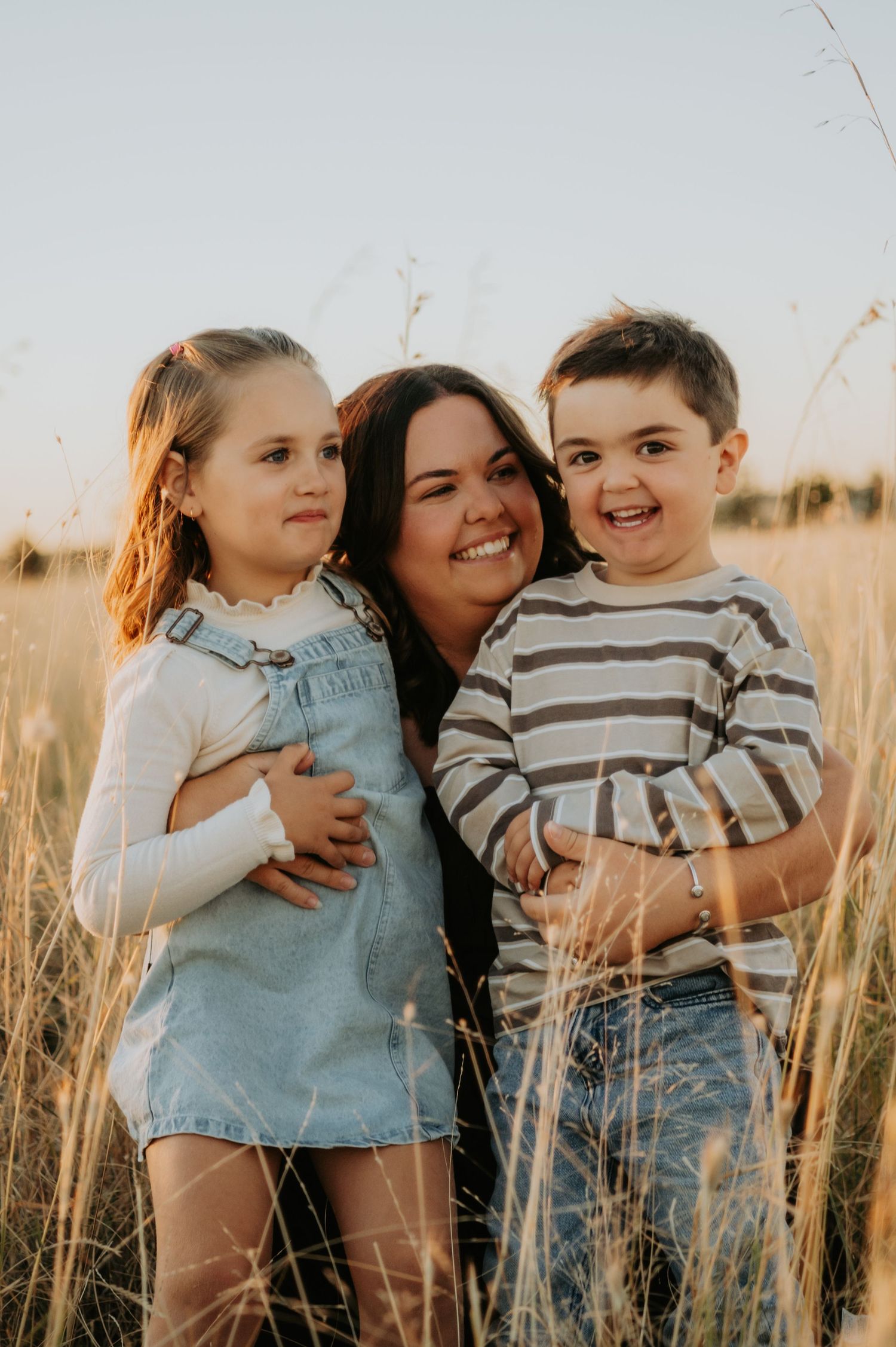 A smiling woman hugs two young children in a golden wheat field during sunset.