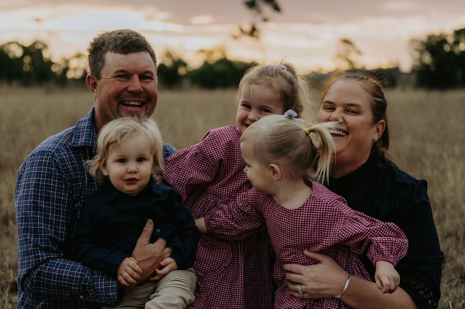 Happy family of five laughing together in a golden field at sunset, parents holding young children.
