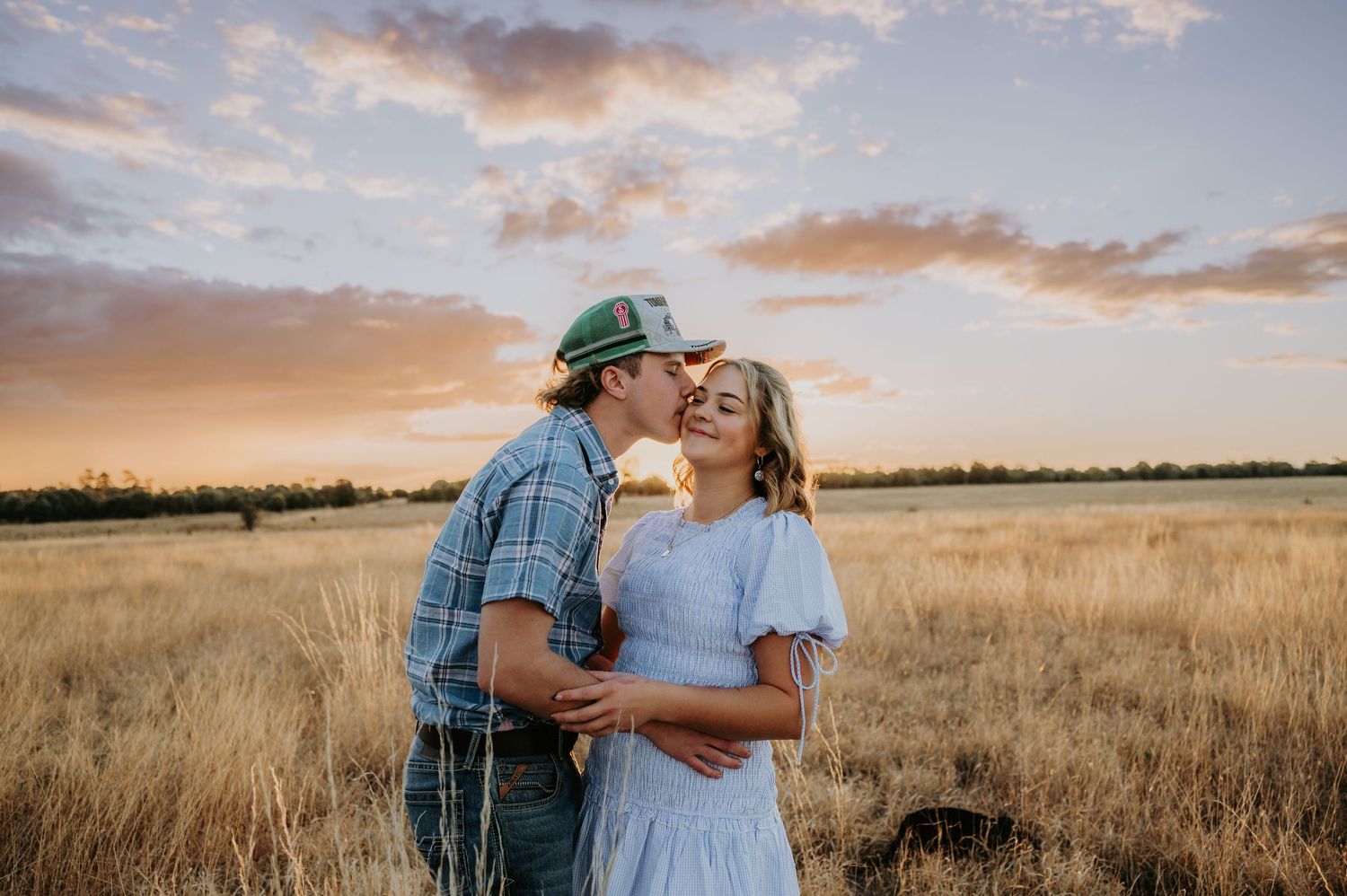 Couple sharing a romantic kiss at sunset in a golden wheat field under a colorful sky.