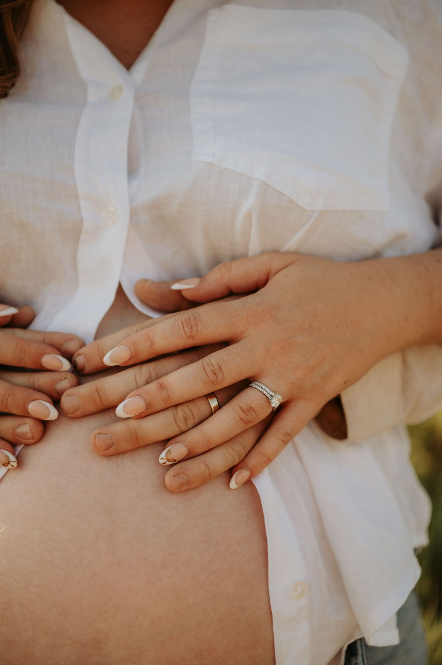 Close-up of hands with wedding rings resting on a pregnant belly against a white shirt.