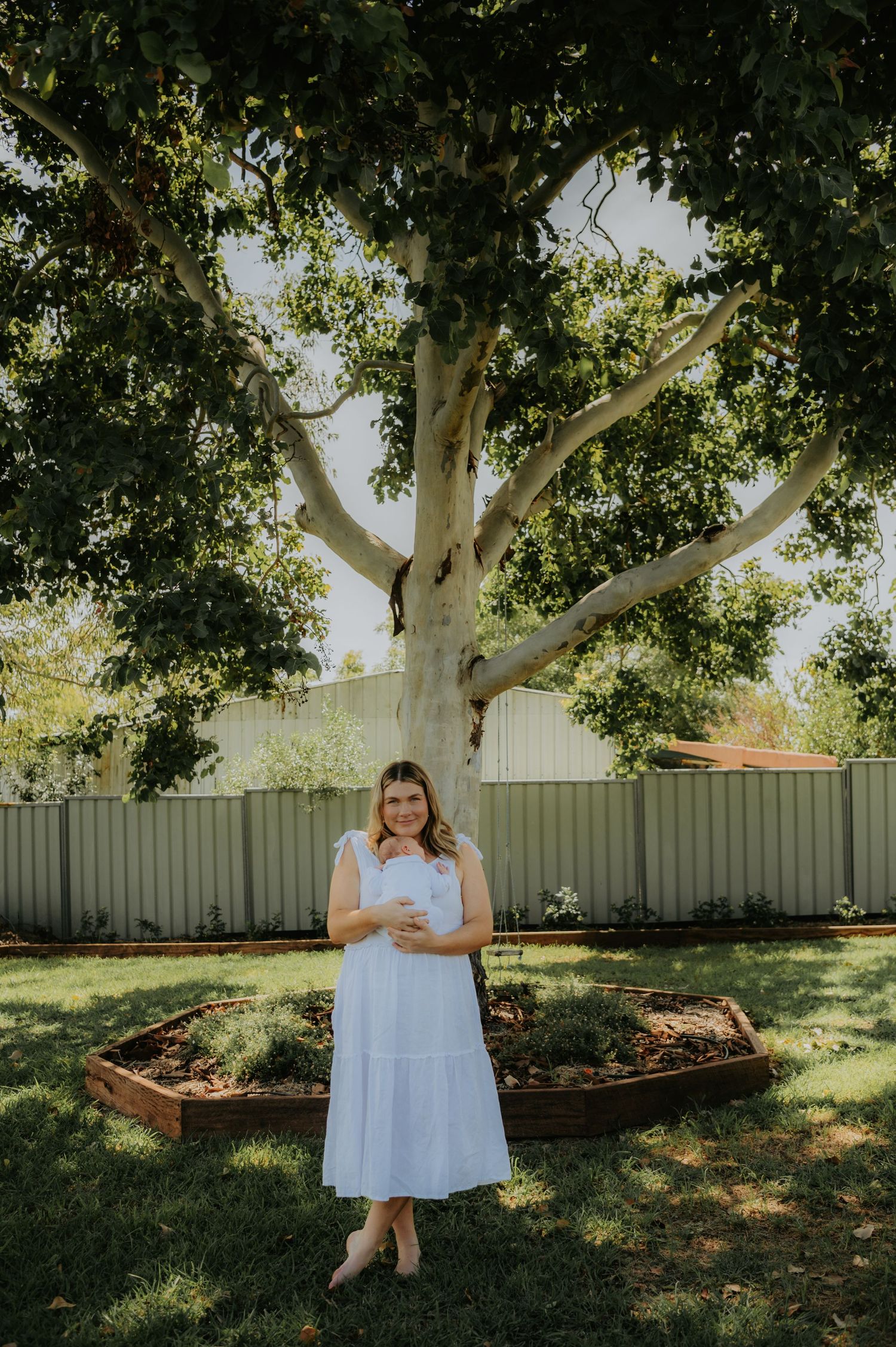 A person in a light blue dress sits under a large shady tree in a backyard garden setting on a sunny day.