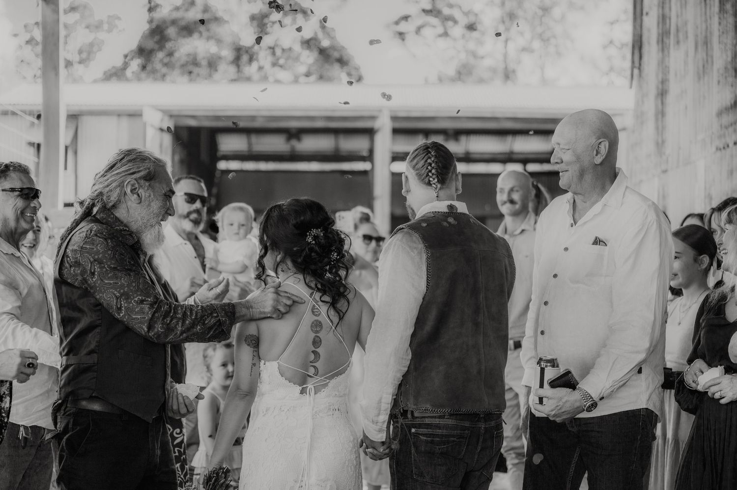 Black and white wedding ceremony photo with bride and groom holding hands surrounded by guests outdoors.