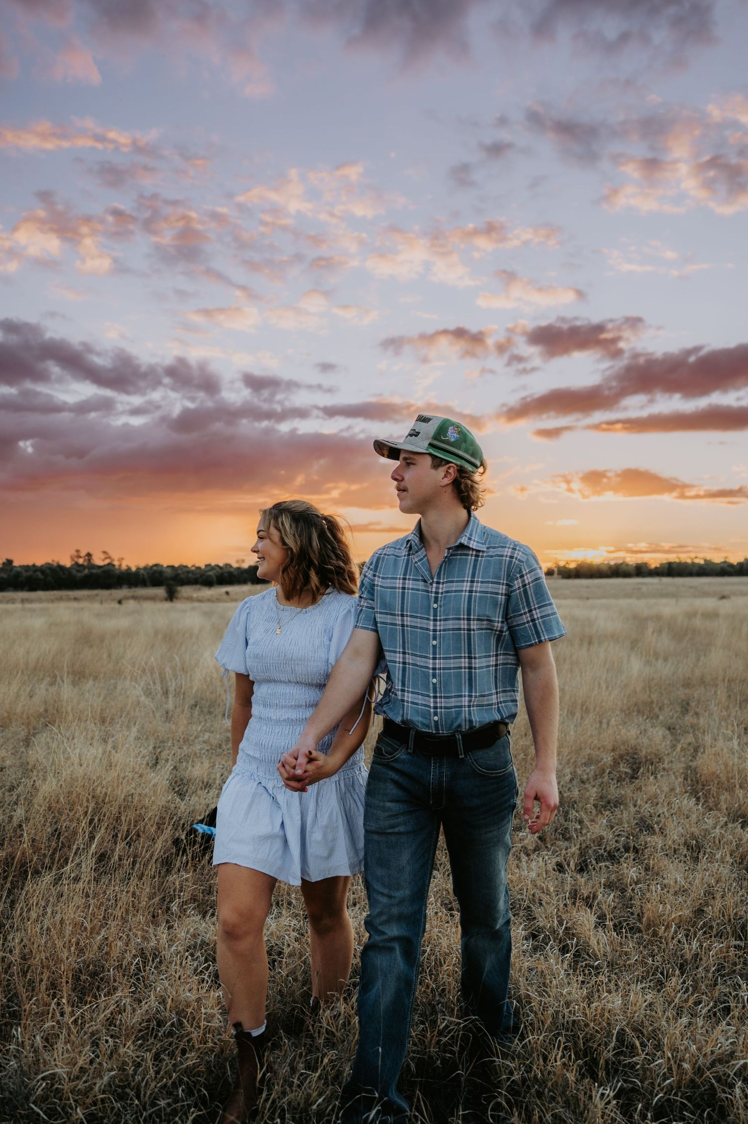 Couple holding hands in golden field at sunset with dramatic pink and purple cloudy sky backdrop.