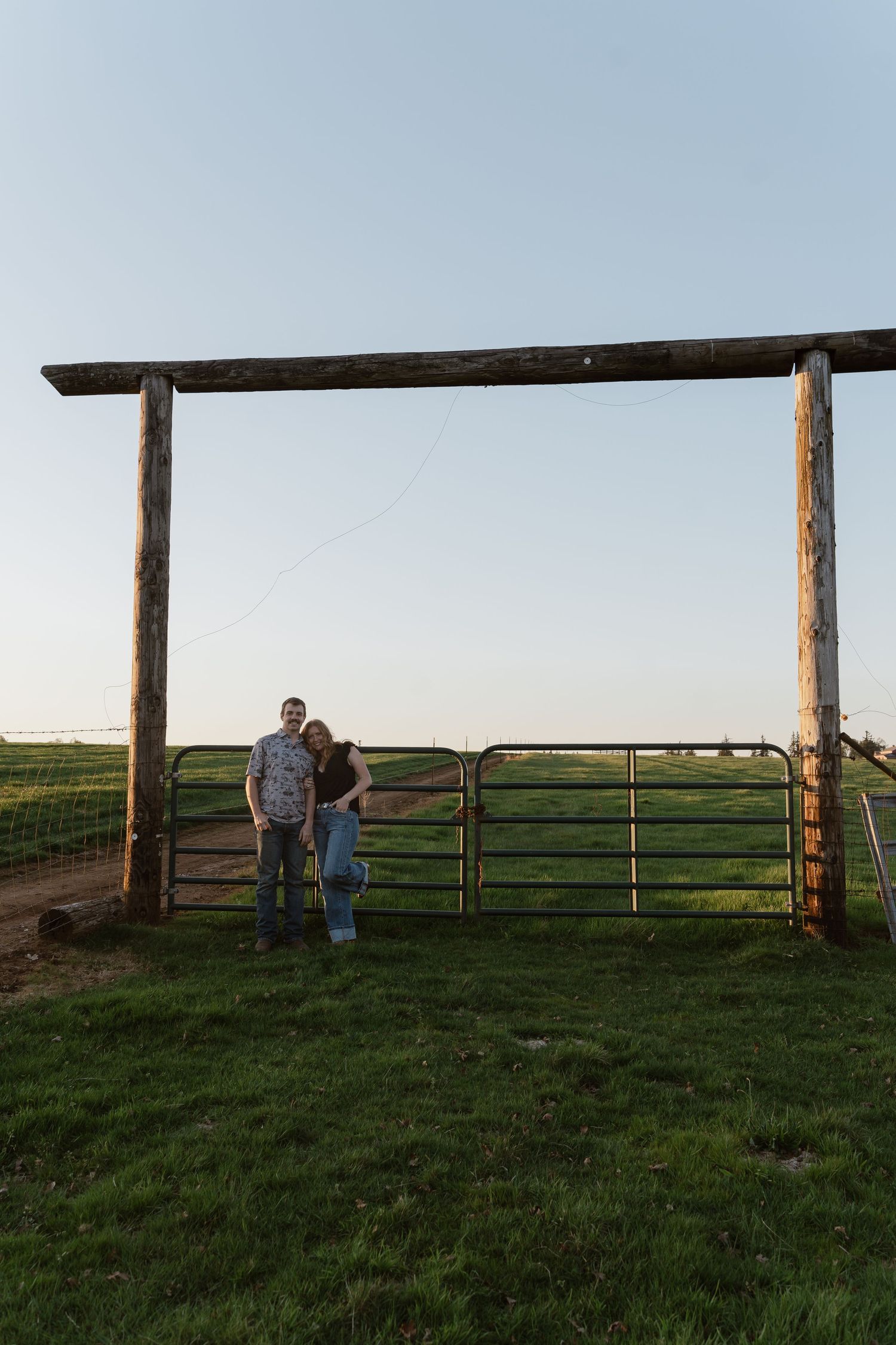 Couple embracing beside a rustic metal farm gate framed by wooden posts in an open green field.