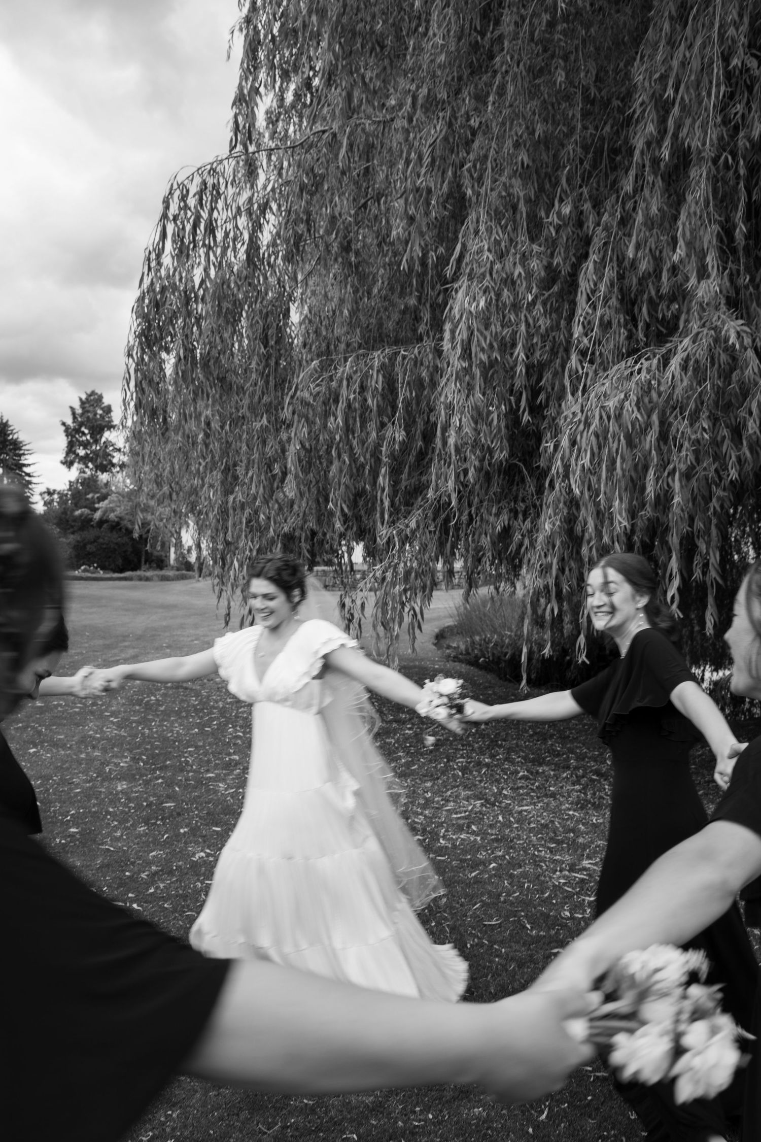 Black and white photo of laughing bride in white gown dancing with bridesmaids under a weeping willow tree.