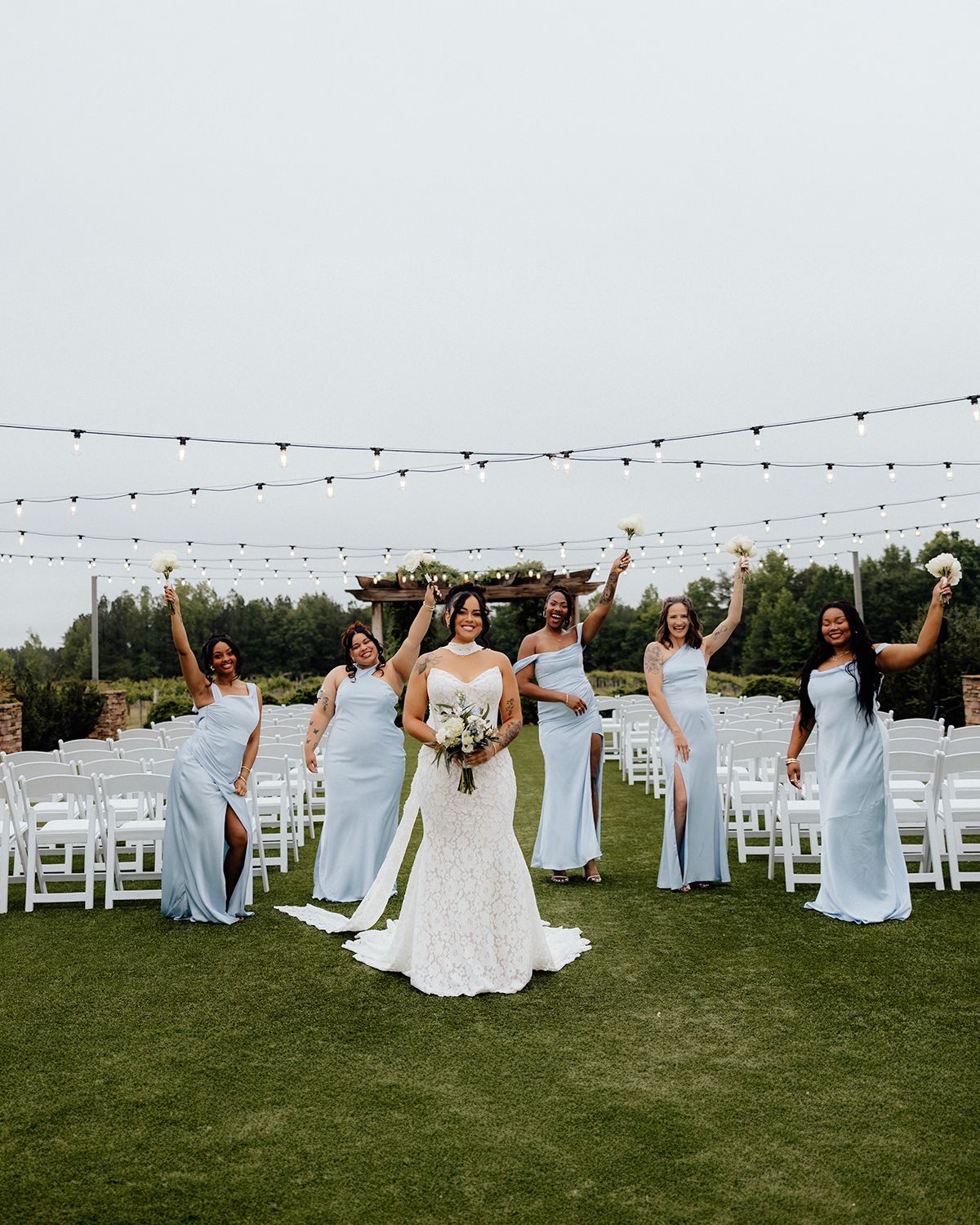 Bride in lace gown poses with cheering bridesmaids in light blue dresses on green lawn at outdoor wedding ceremony.