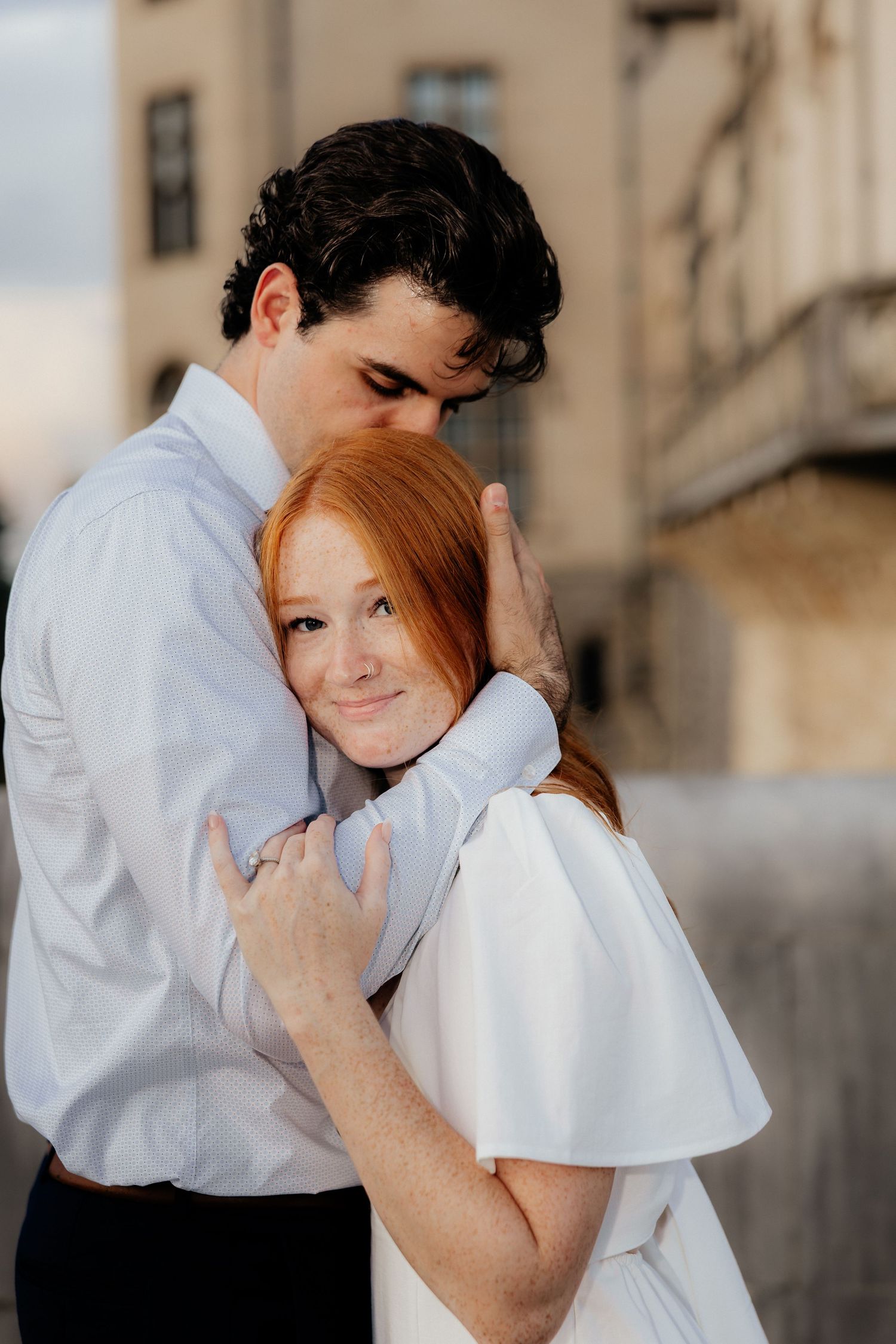 A couple embraces tenderly, the redheaded woman in white smiling as the dark-haired man holds her close outdoors.