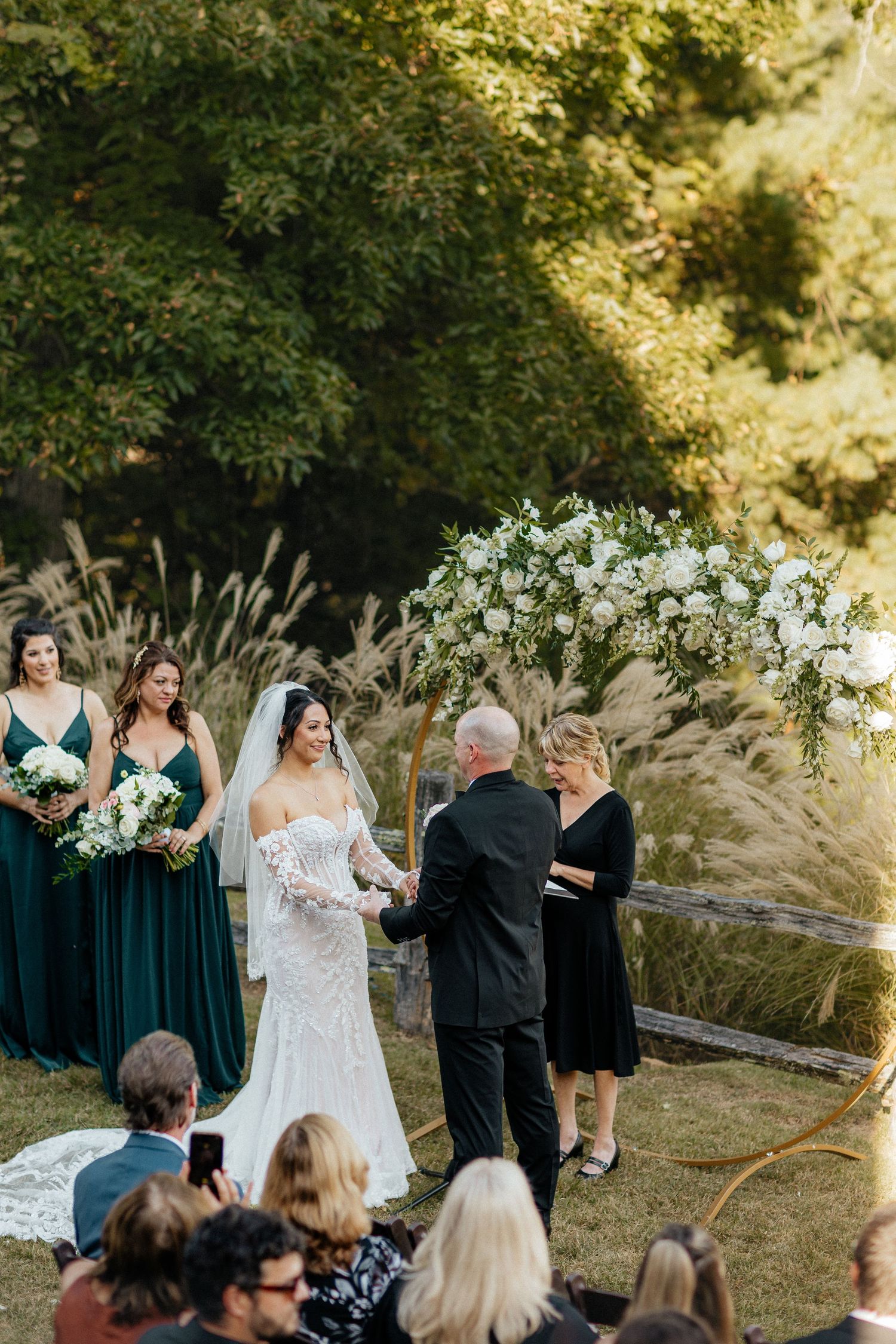 Outdoor wedding ceremony with bride in lace gown, bridesmaids in emerald dresses, and white floral arch amid pampas grass.