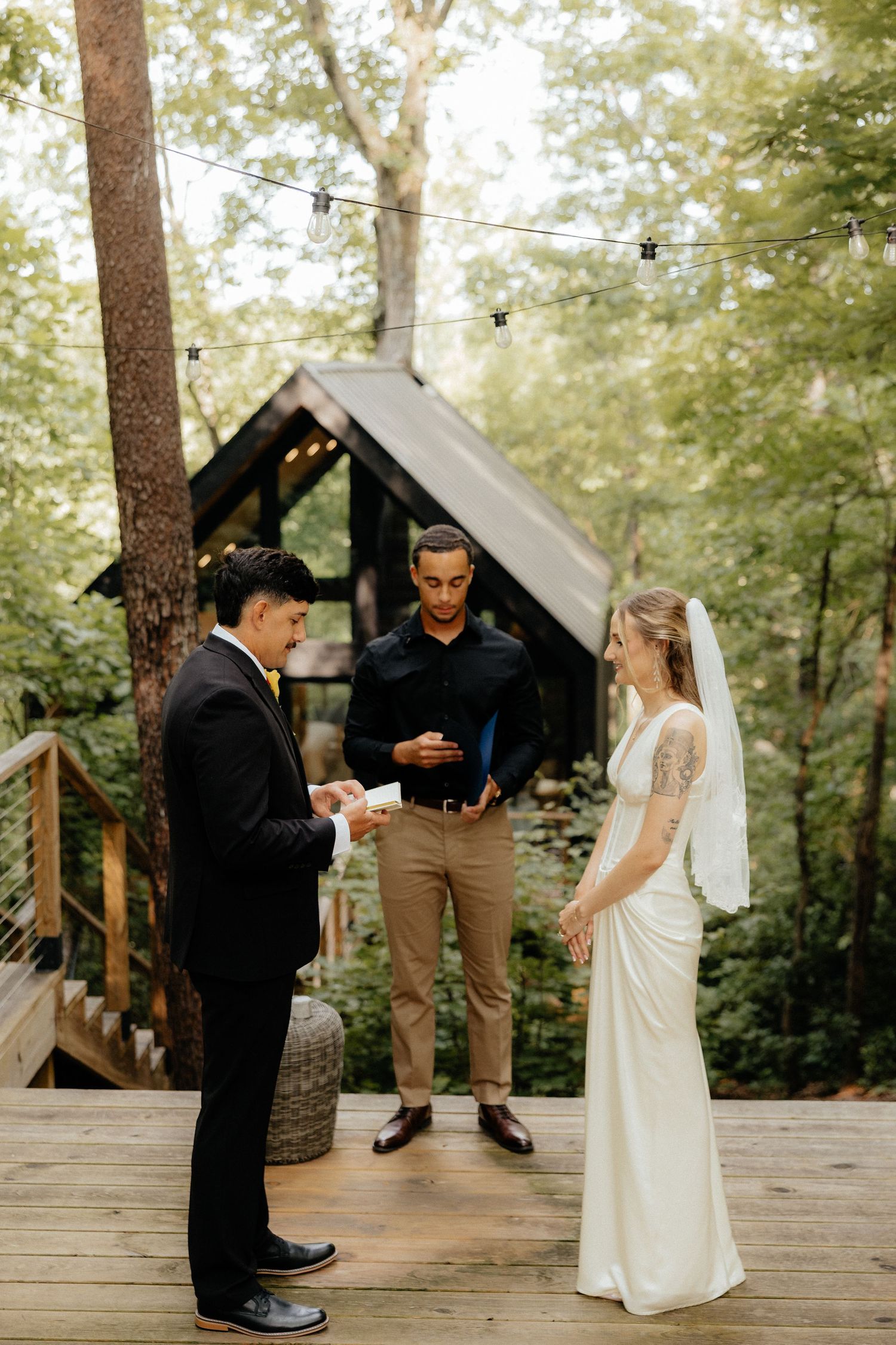 Bride in a white gown and veil exchanges vows with a groom in a black suit as an officiant looks on during an outdoor forest wedding.