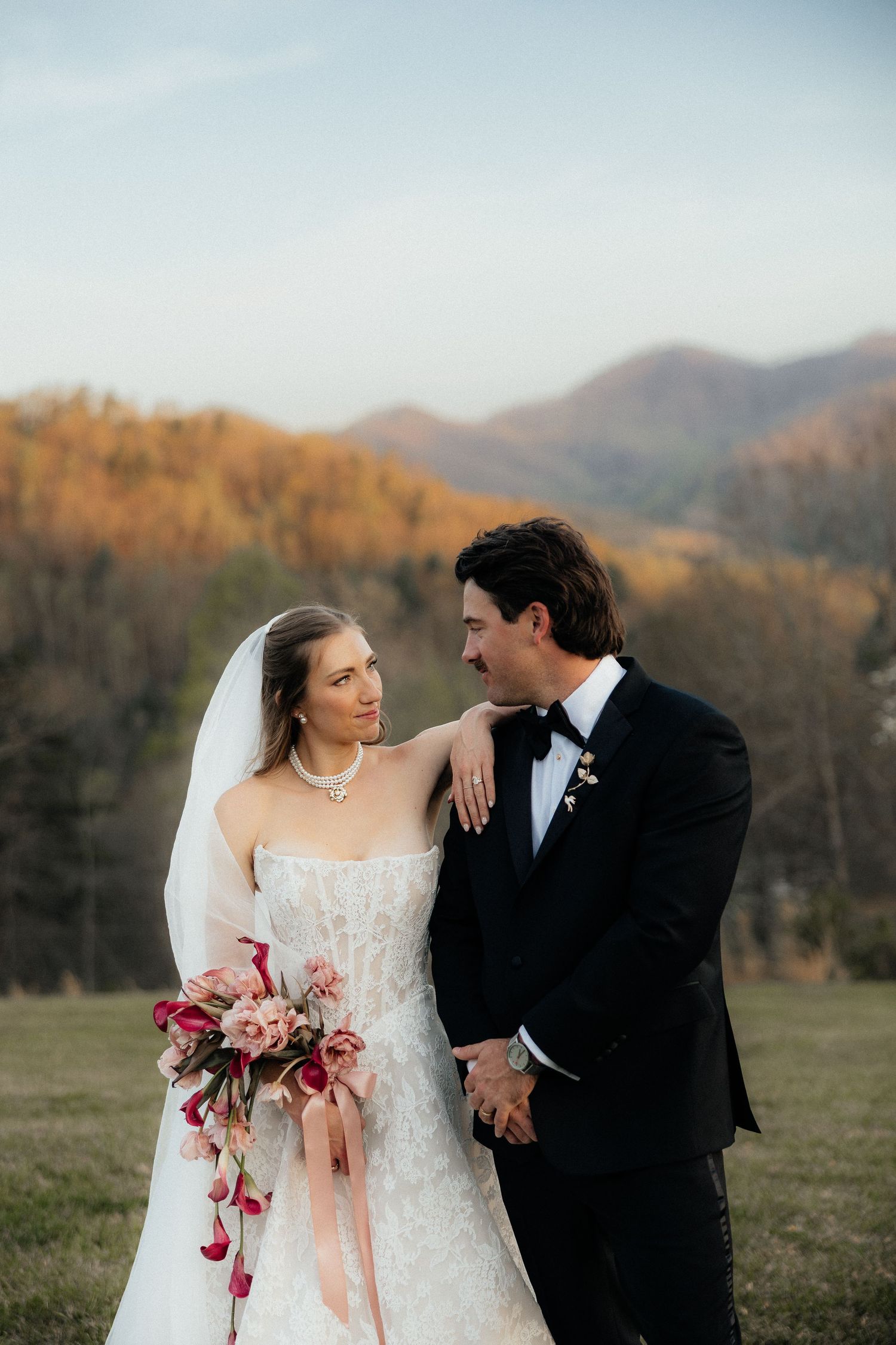 Bride in a lace gown and veil holding a pink bouquet embraces her groom in a black tuxedo against autumn mountains at sunset.