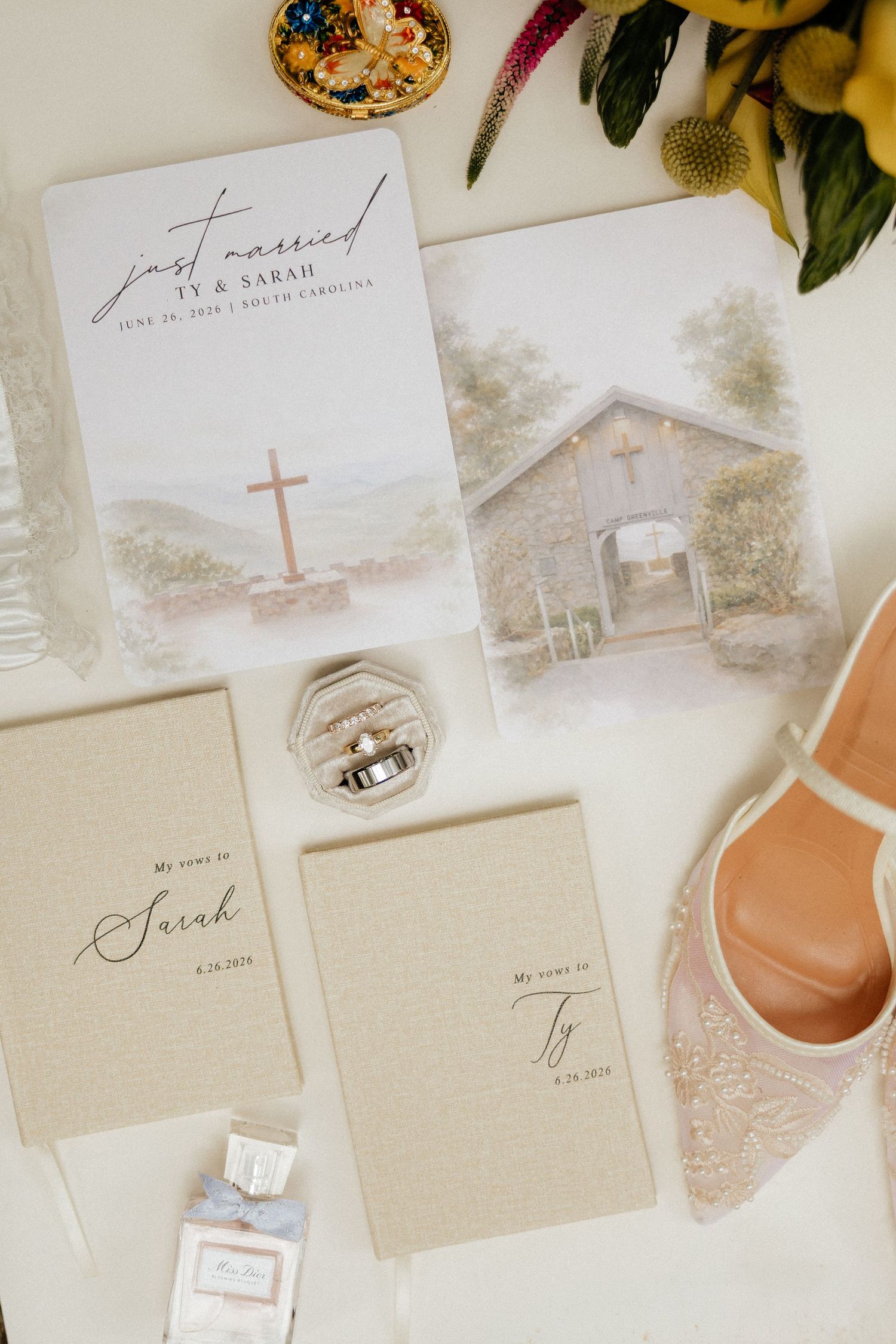 Wedding flat lay with Ty and Sarah's just married card, vow books, ring box, lace heels and a watercolor chapel print.