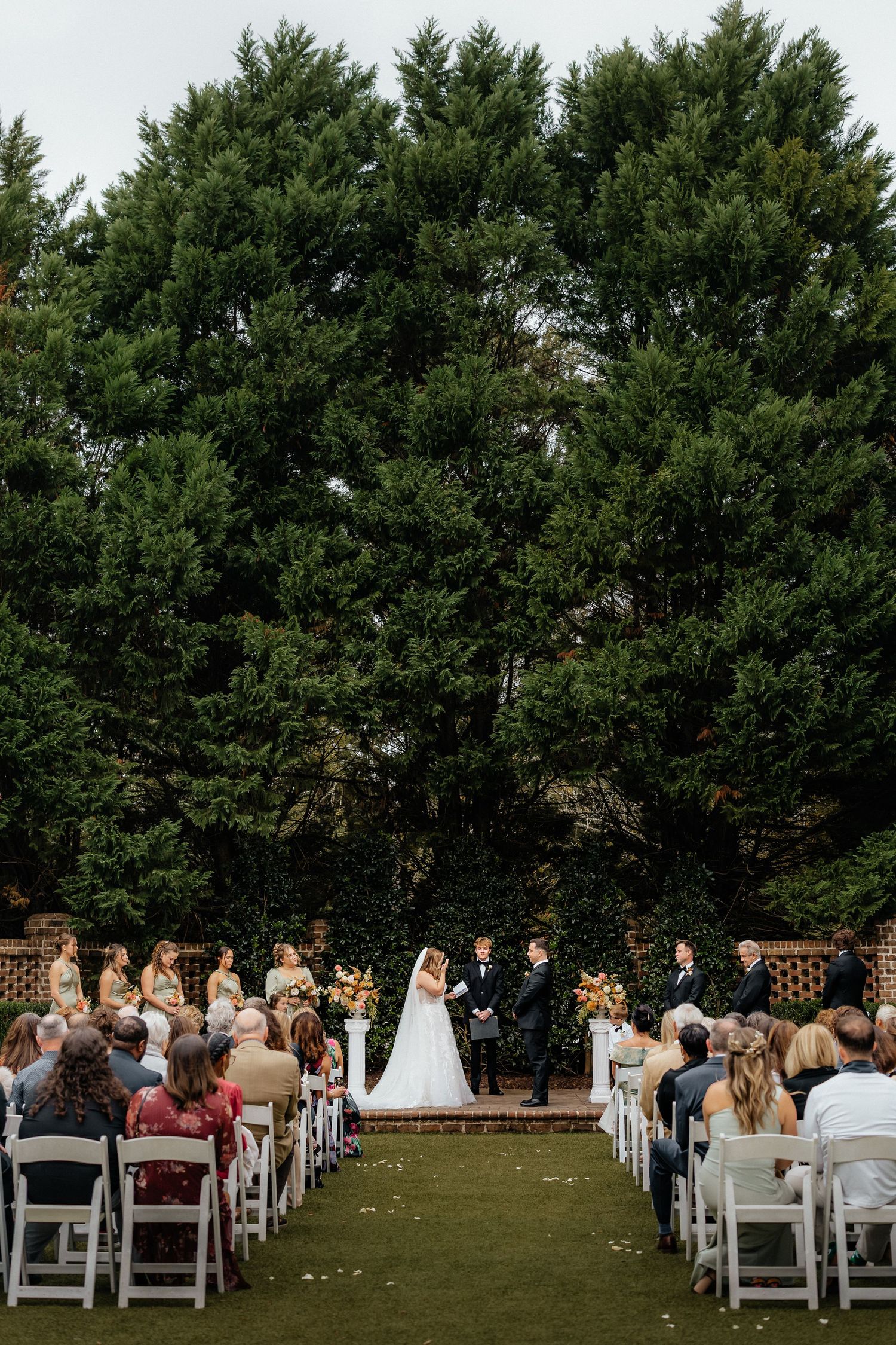 An outdoor wedding ceremony takes place on a lawn with white chairs and tall evergreen trees creating a natural backdrop.