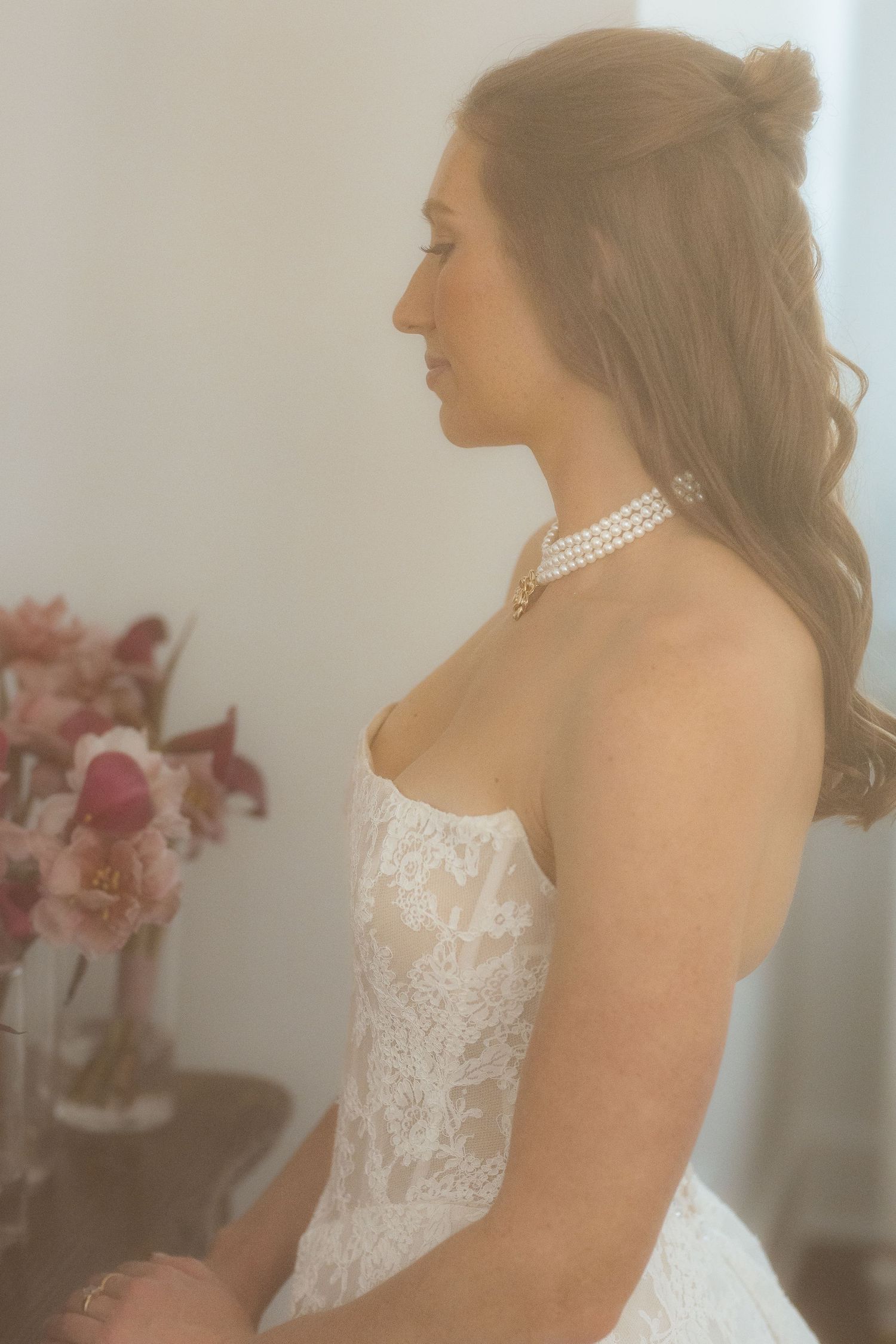 Bride in strapless lace wedding gown with pearl choker necklace, sitting in profile view with pink flowers in background.