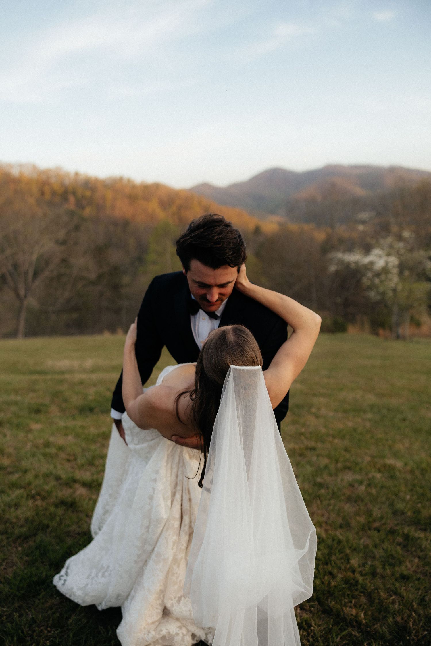 Bride and groom share a romantic dip kiss in a green field with mountain backdrop at golden hour.