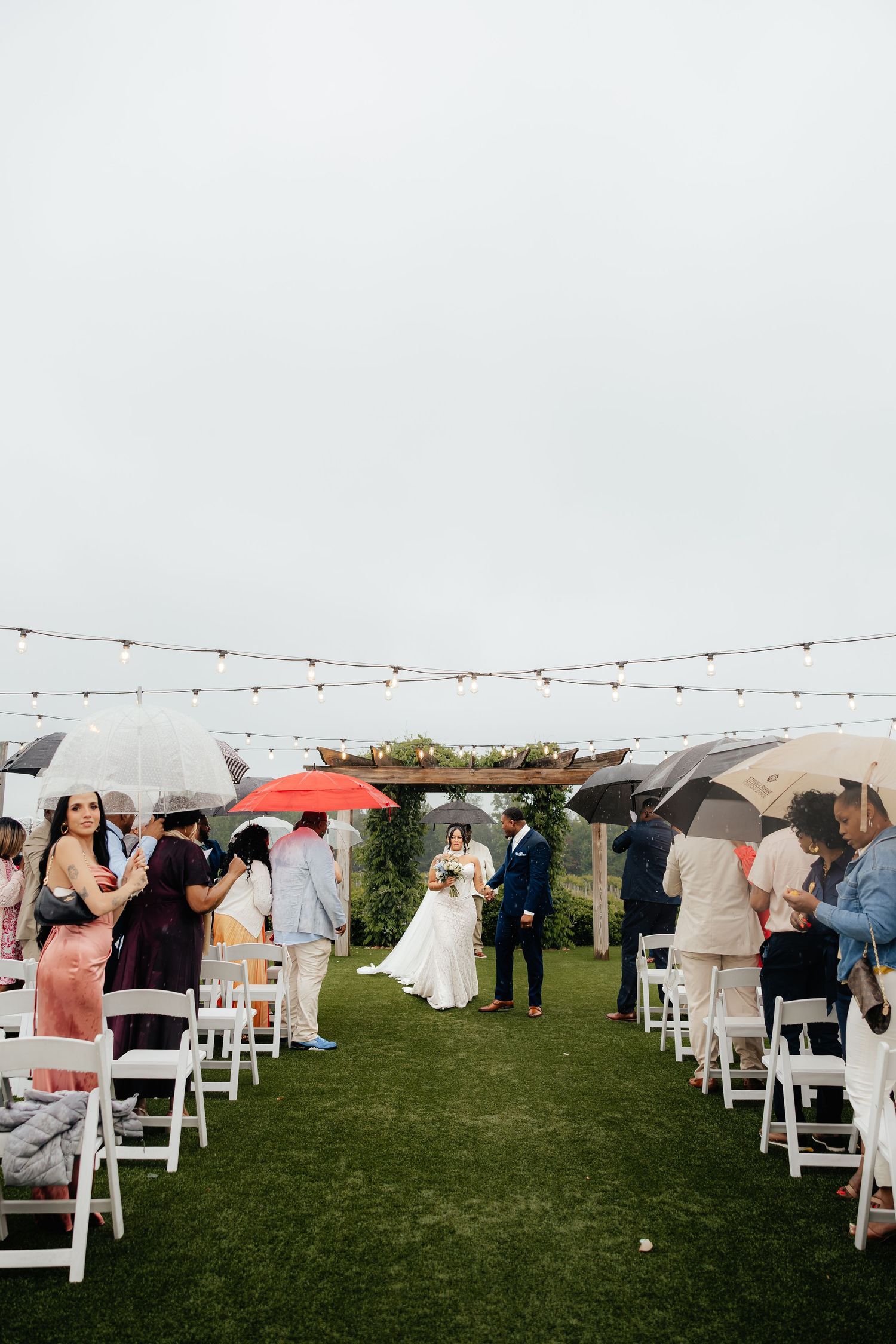 Outdoor wedding ceremony on green lawn with guests holding umbrellas in rain, string lights overhead, bride and groom at altar.