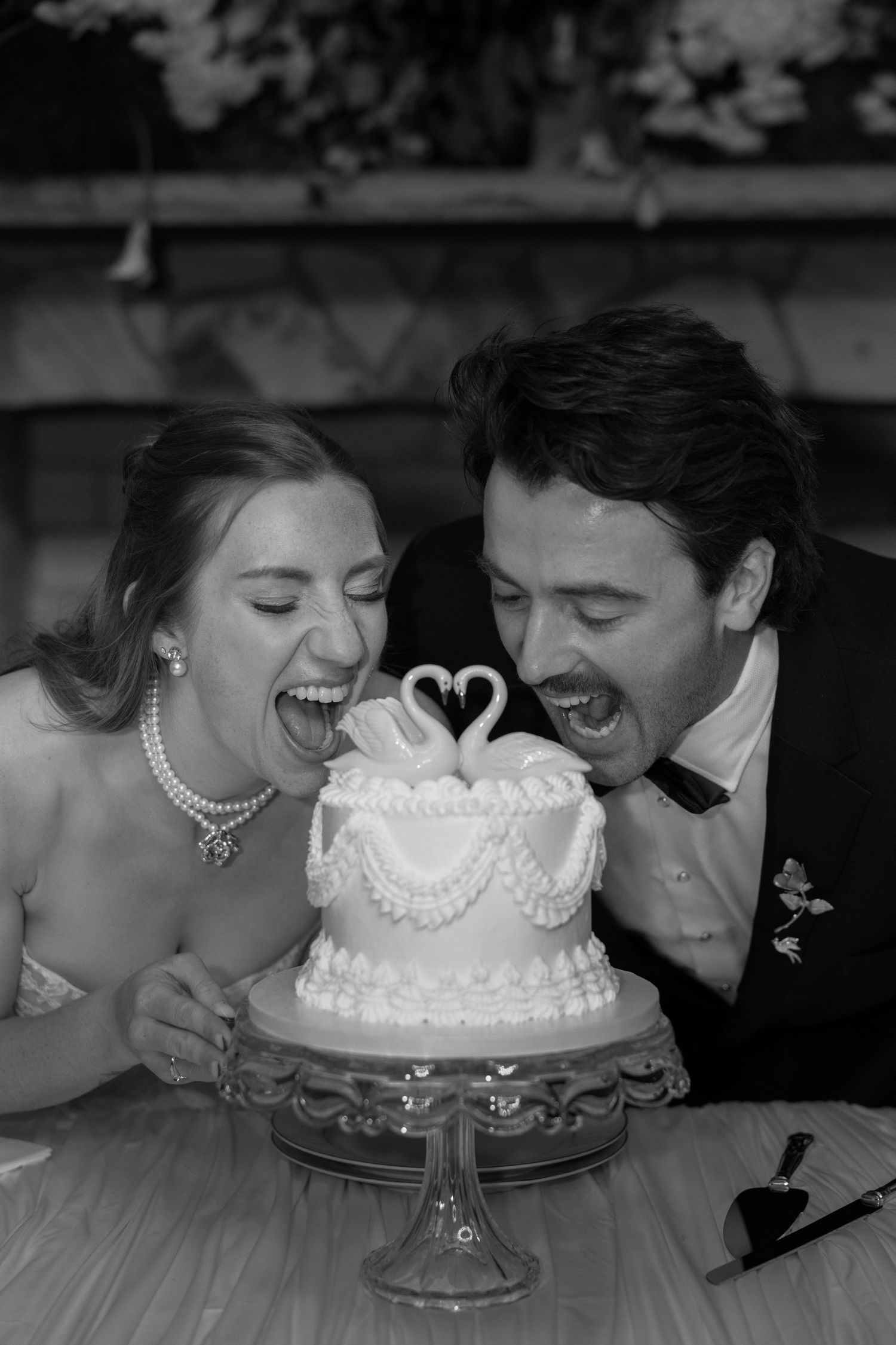 Black and white photo of a bride and groom laughing with mouths open, about to bite a small wedding cake topped with swans.