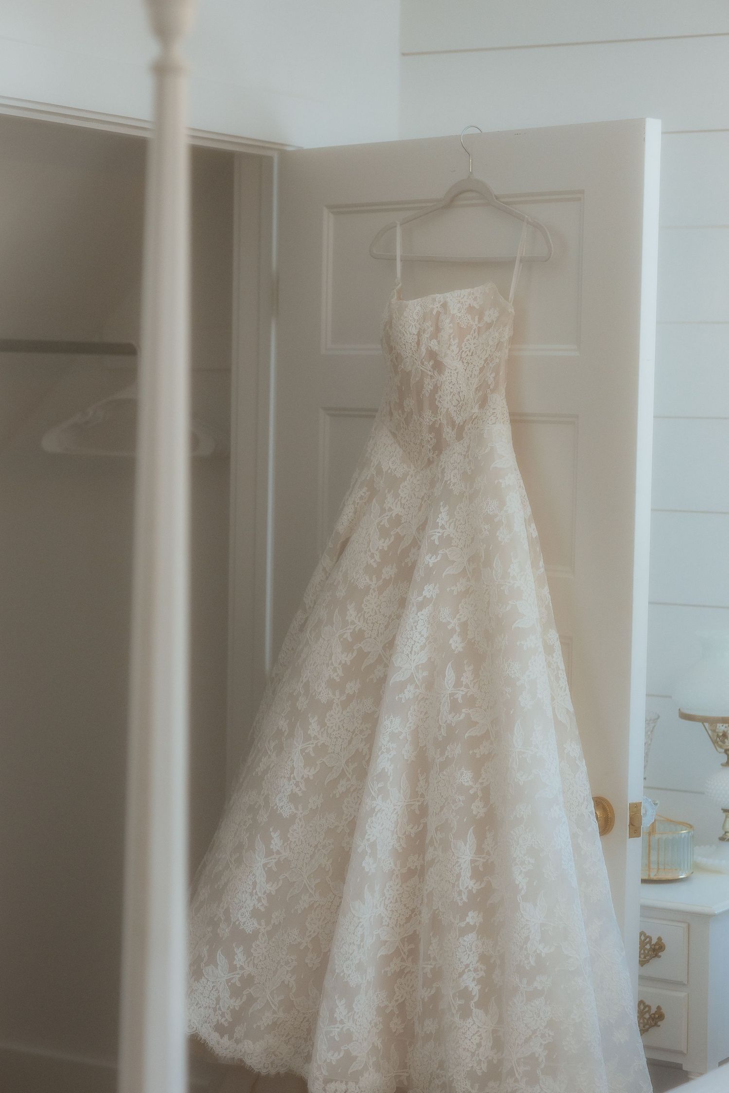 Elegant strapless lace ball gown wedding dress hanging on a door hanger in a bright, softly lit bridal suite.