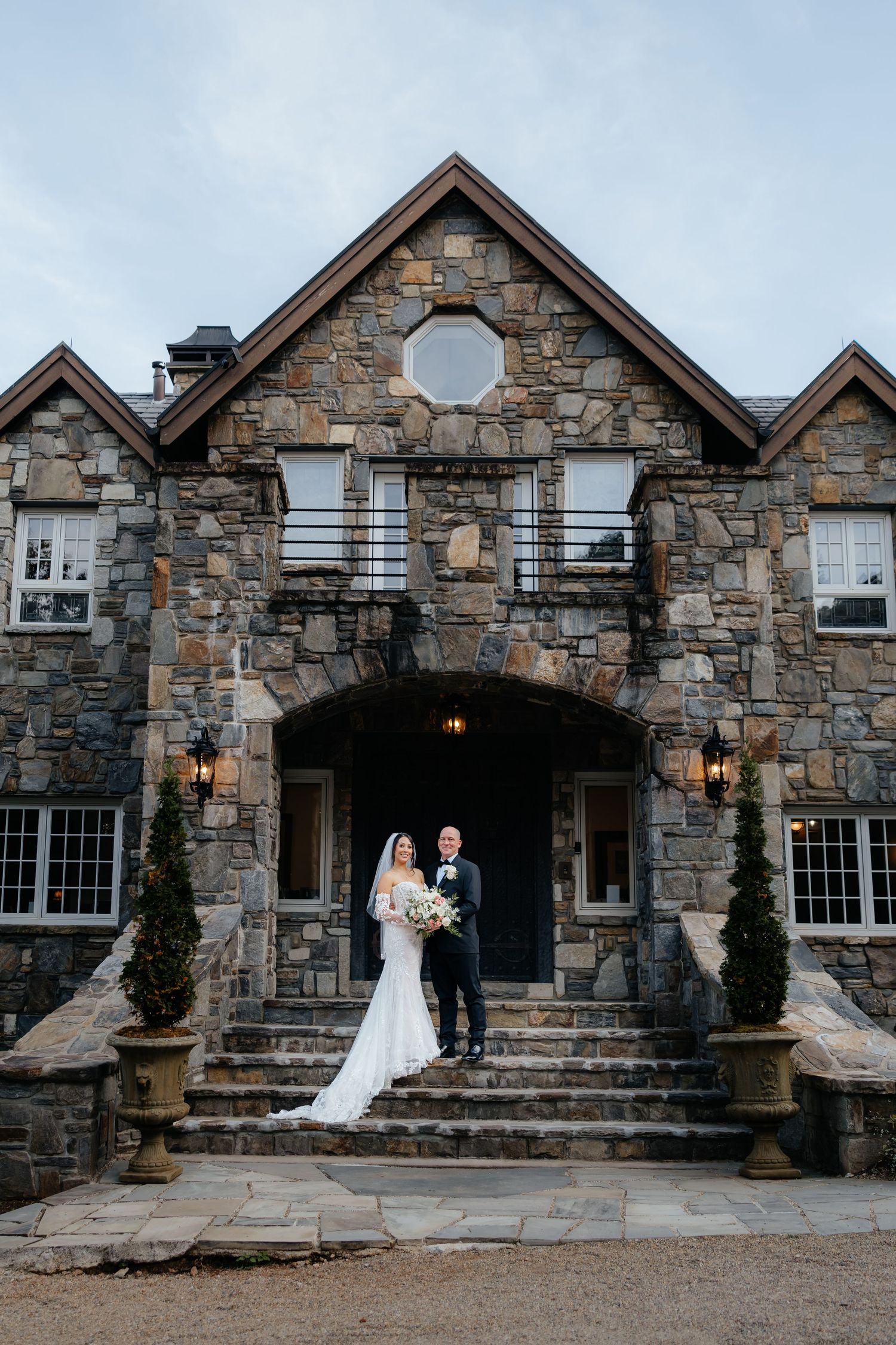 Bride in a white gown holding a bouquet and groom in a dark suit pose on the stone steps of a rustic stone mansion wedding venue.