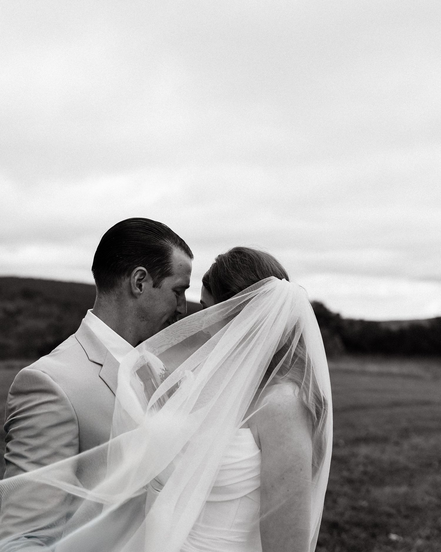 Black and white wedding photo of bride and groom kissing under flowing veil in an open field.