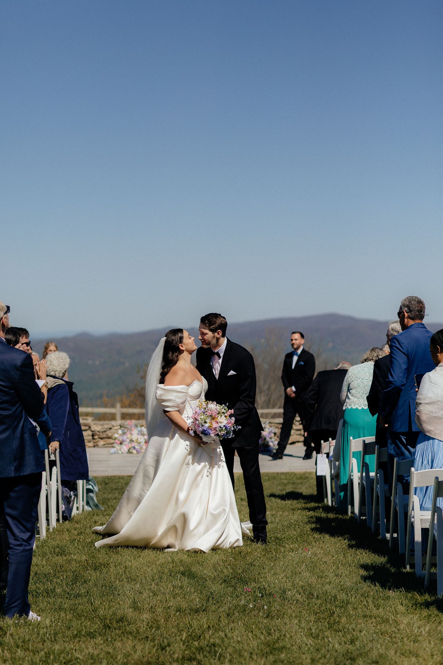 Bride and groom share a kiss at outdoor wedding ceremony with mountain views and guests seated along the aisle.