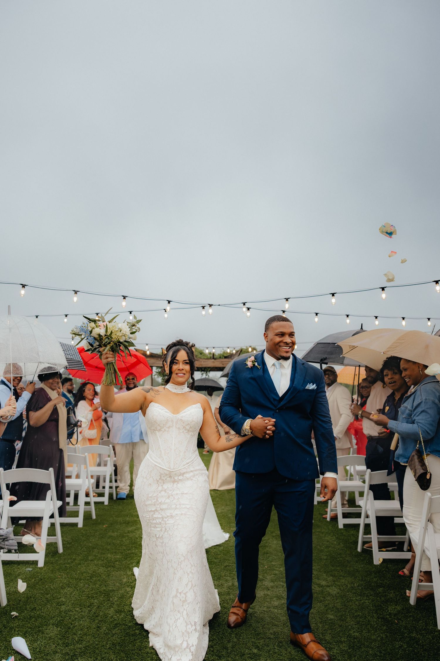 Newlywed couple walks joyfully down the aisle after outdoor wedding ceremony as guests celebrate with umbrellas.