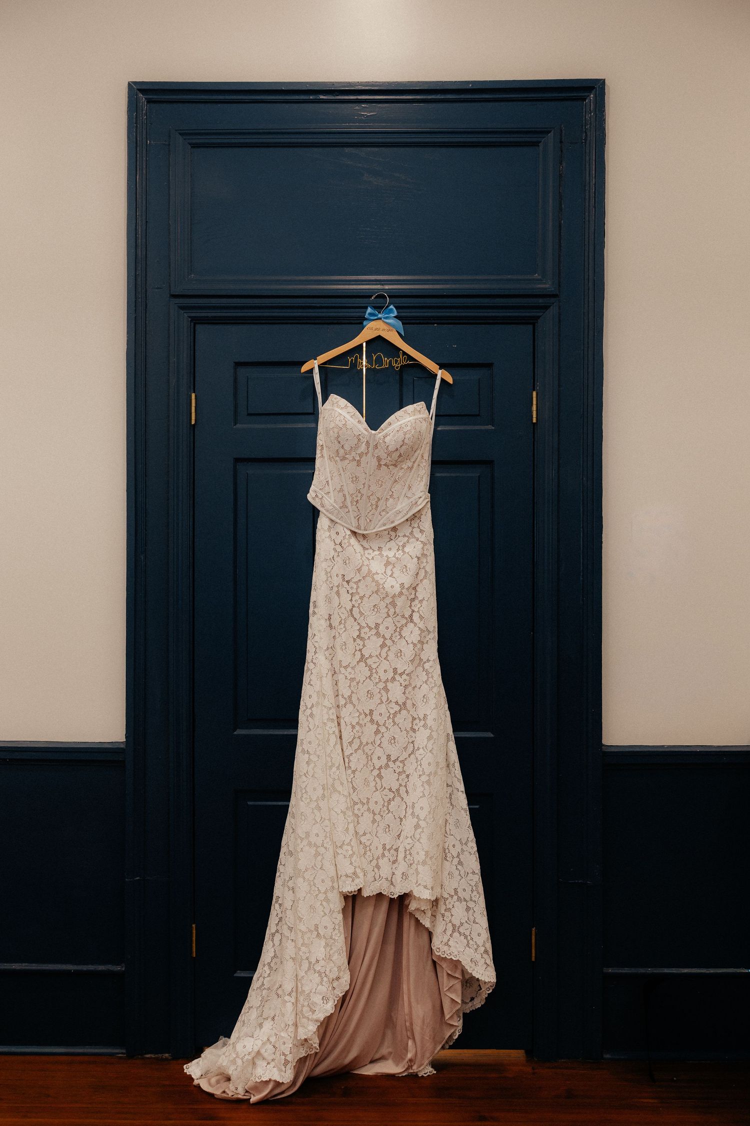 Elegant lace wedding dress hanging on a wooden hanger against a navy blue paneled door in a bridal suite.