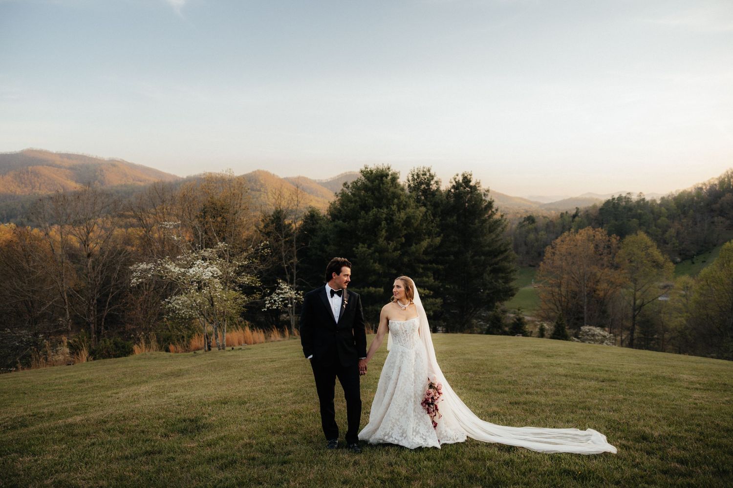 Bride and groom hold hands on a lush green hillside with scenic mountain views and colorful autumn foliage at sunset.
