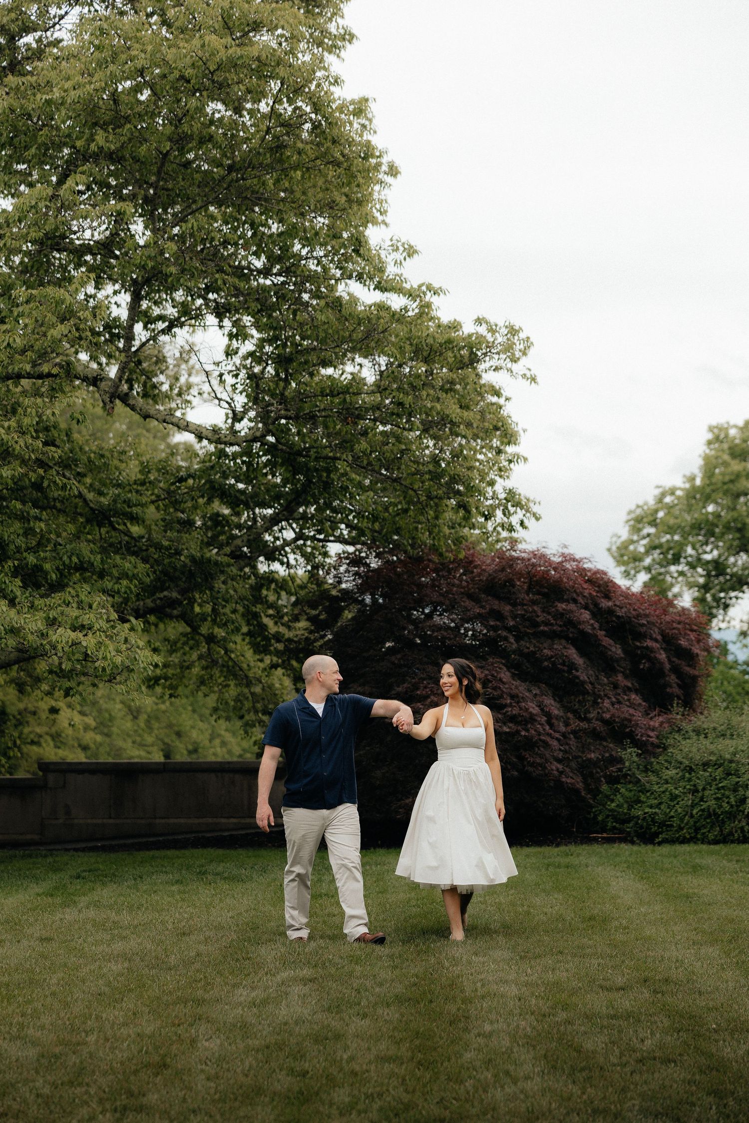 Couple holding hands and dancing on green lawn surrounded by lush trees in an outdoor garden setting.