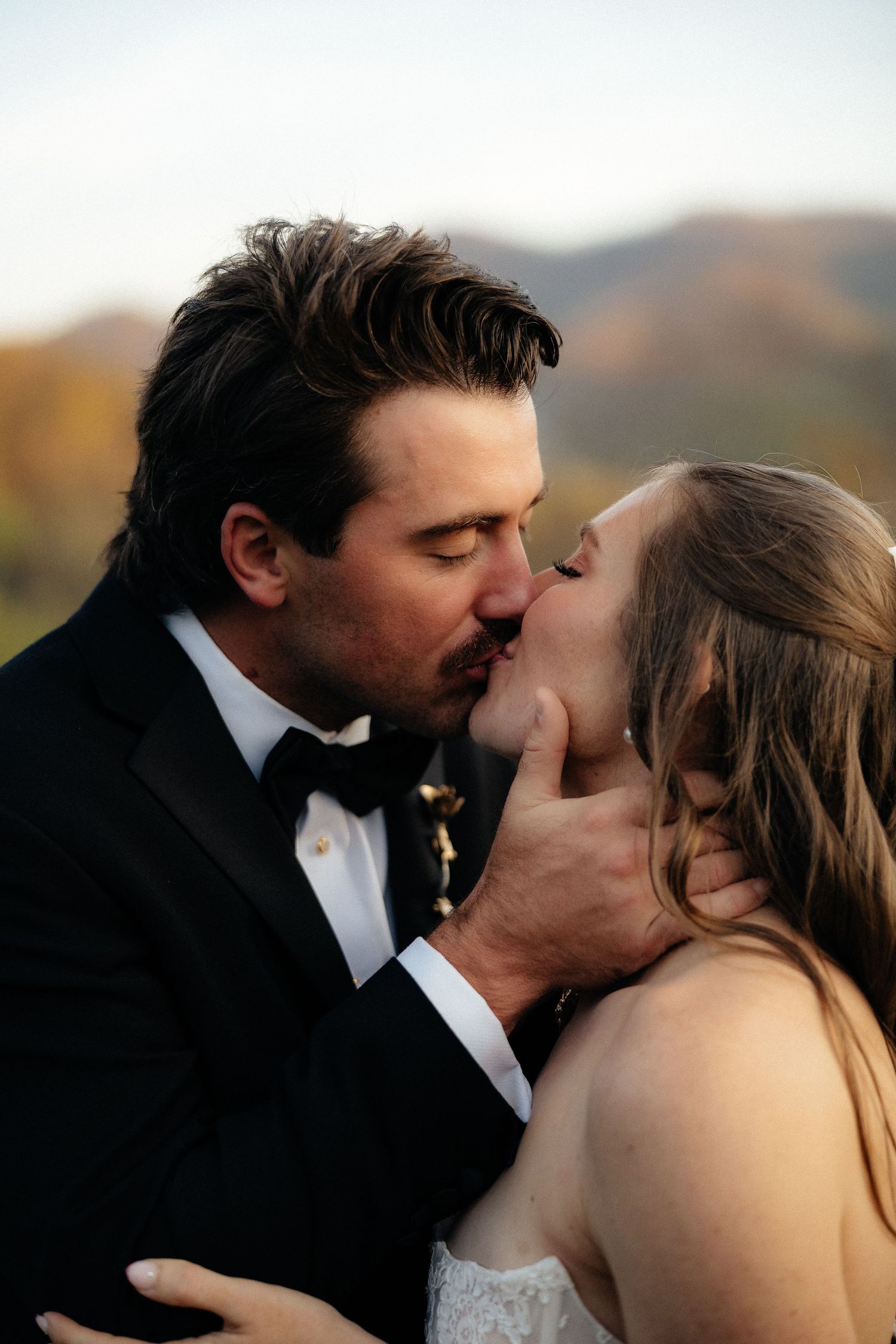 Groom in a black tuxedo and bow tie gently cradles his bride's chin as they kiss against a blurred autumn mountain backdrop.