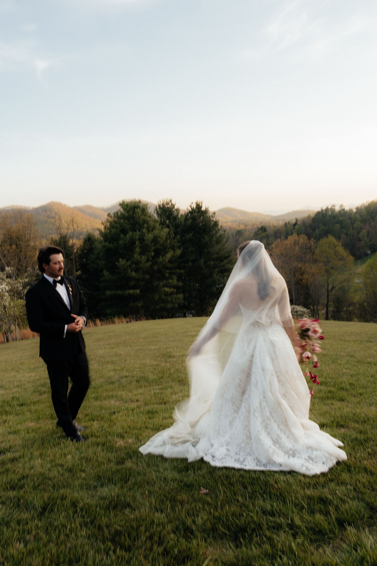 Bride in a lace gown and veil holding a cascading bouquet faces a groom in a black tuxedo on a grassy hilltop with mountain views.