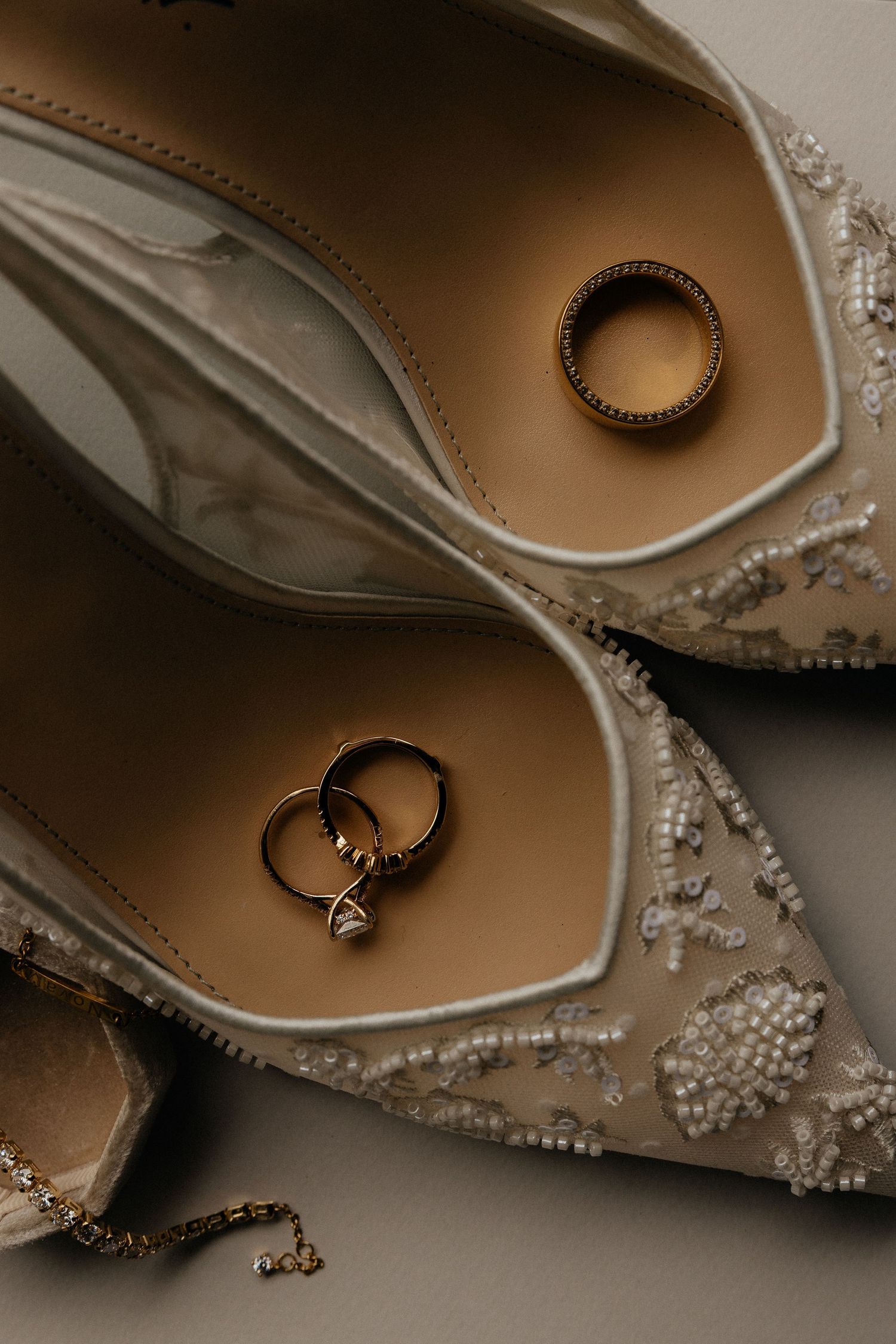 Elegant beaded bridal heels with gold wedding rings and diamond engagement ring nestled inside, styled on a neutral background.