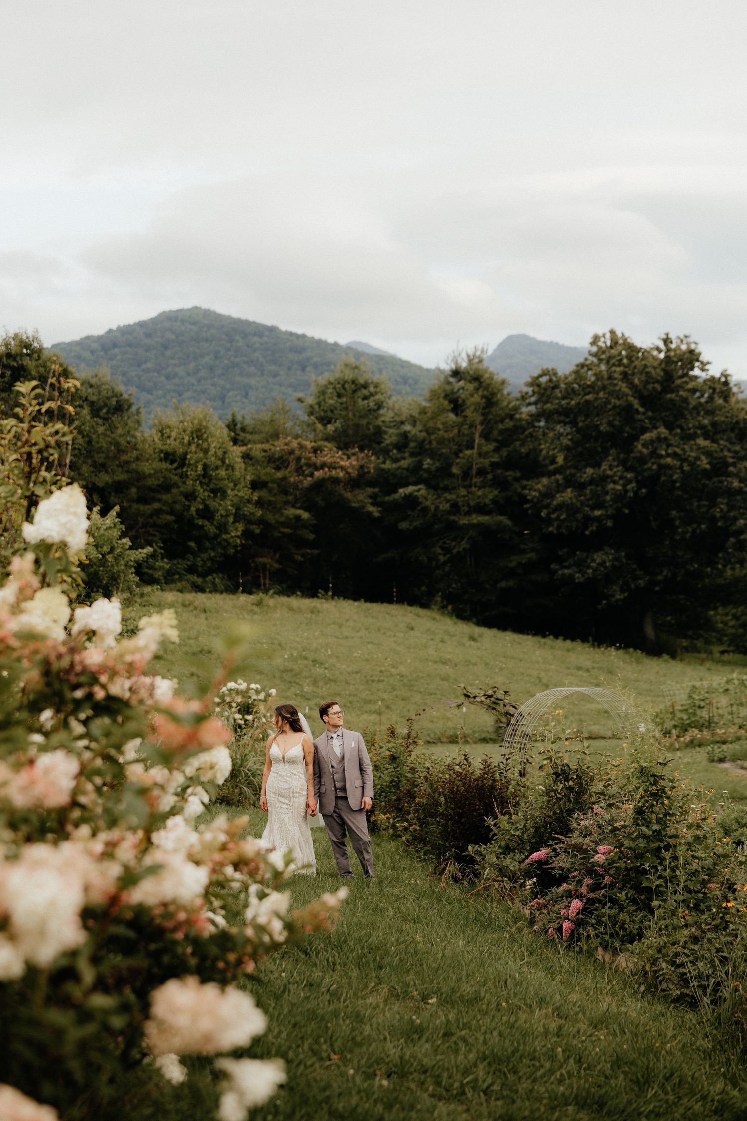 Bride and groom walk through lush green meadow with mountain backdrop and blooming flowers at outdoor wedding.