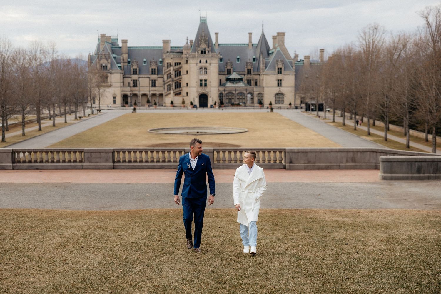 Two figures walk together on a grand estate terrace with stone balustrades and winter trees in the background.