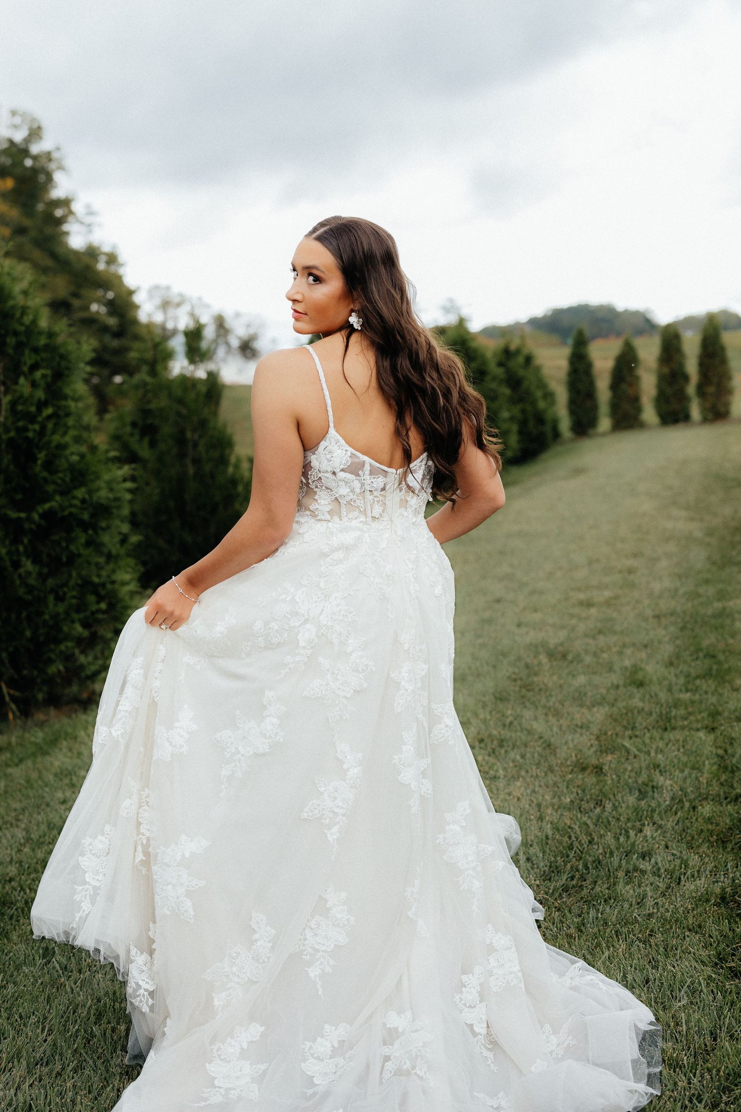 A bride in a flowing white lace wedding dress twirls gracefully in a green hillside meadow on an overcast day.