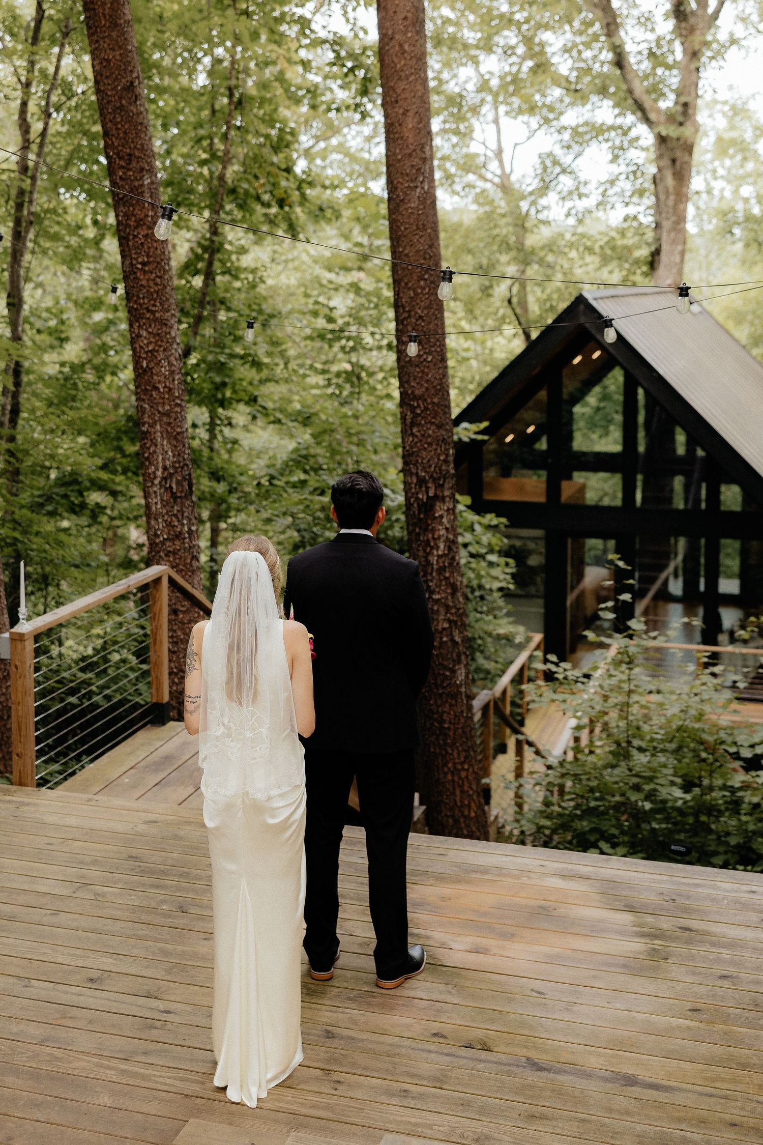 Bride in a white gown and long veil stands beside a groom in a dark suit on a wooden deck in a forest, facing a modern timber cabin.