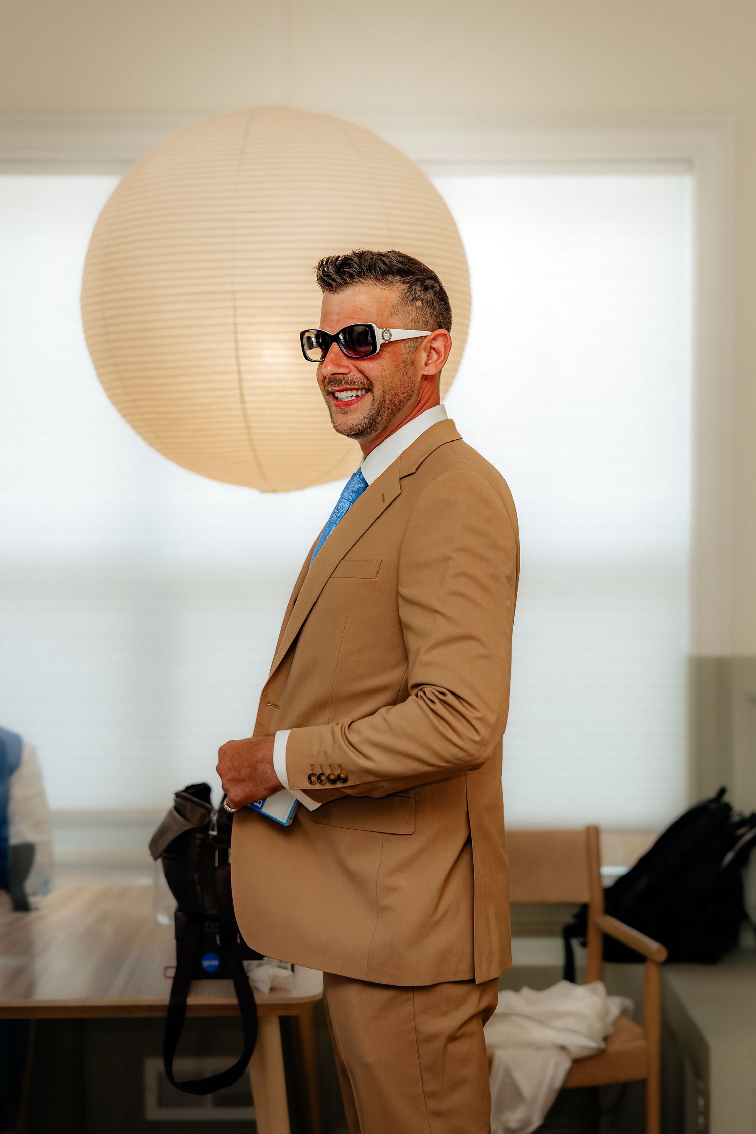 Smiling man in tan suit, blue tie, and sunglasses indoors with a round paper lantern light.