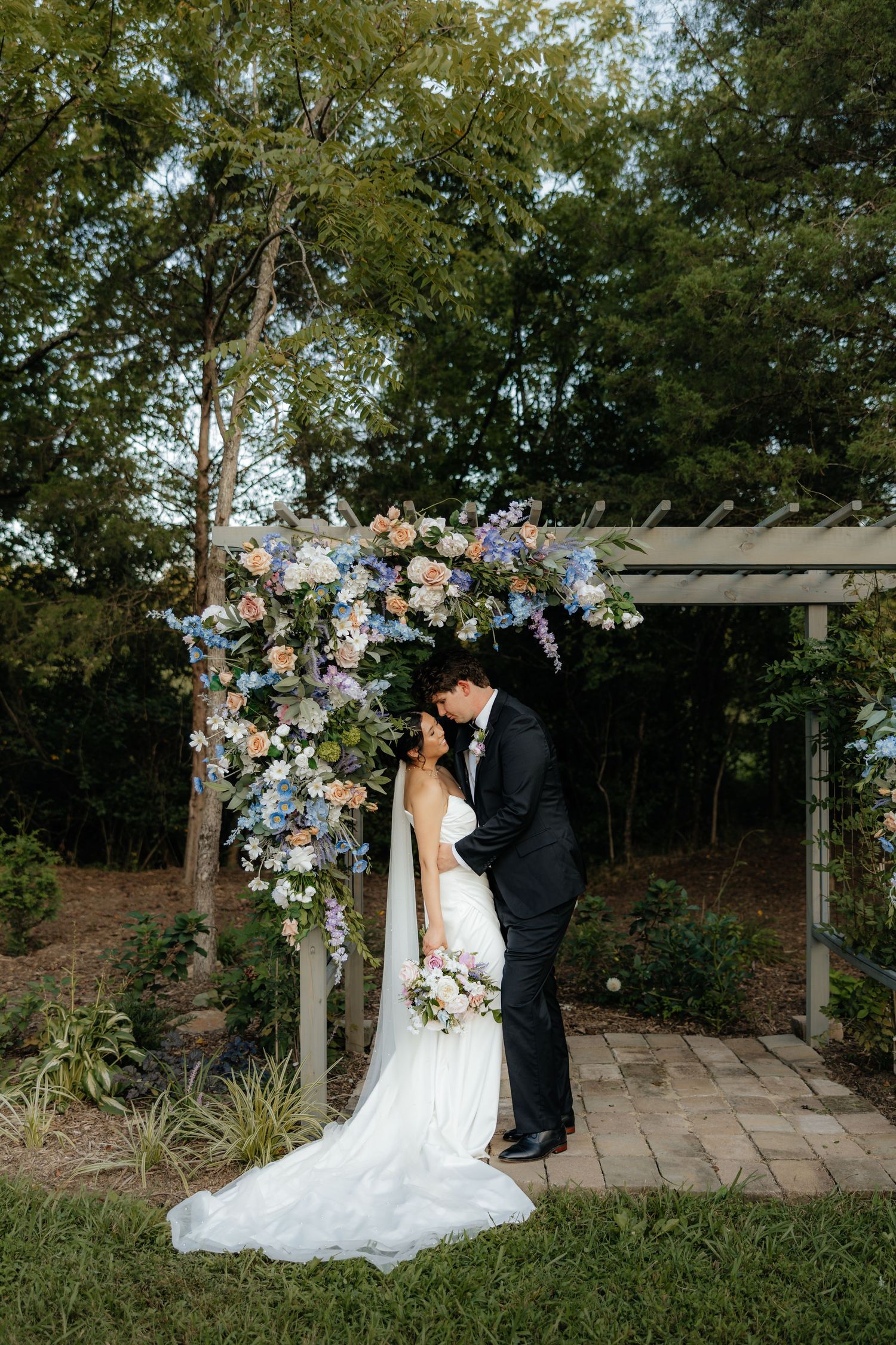 A romantic wedding scene under a wooden pergola decorated with pastel floral arrangements in a garden setting.