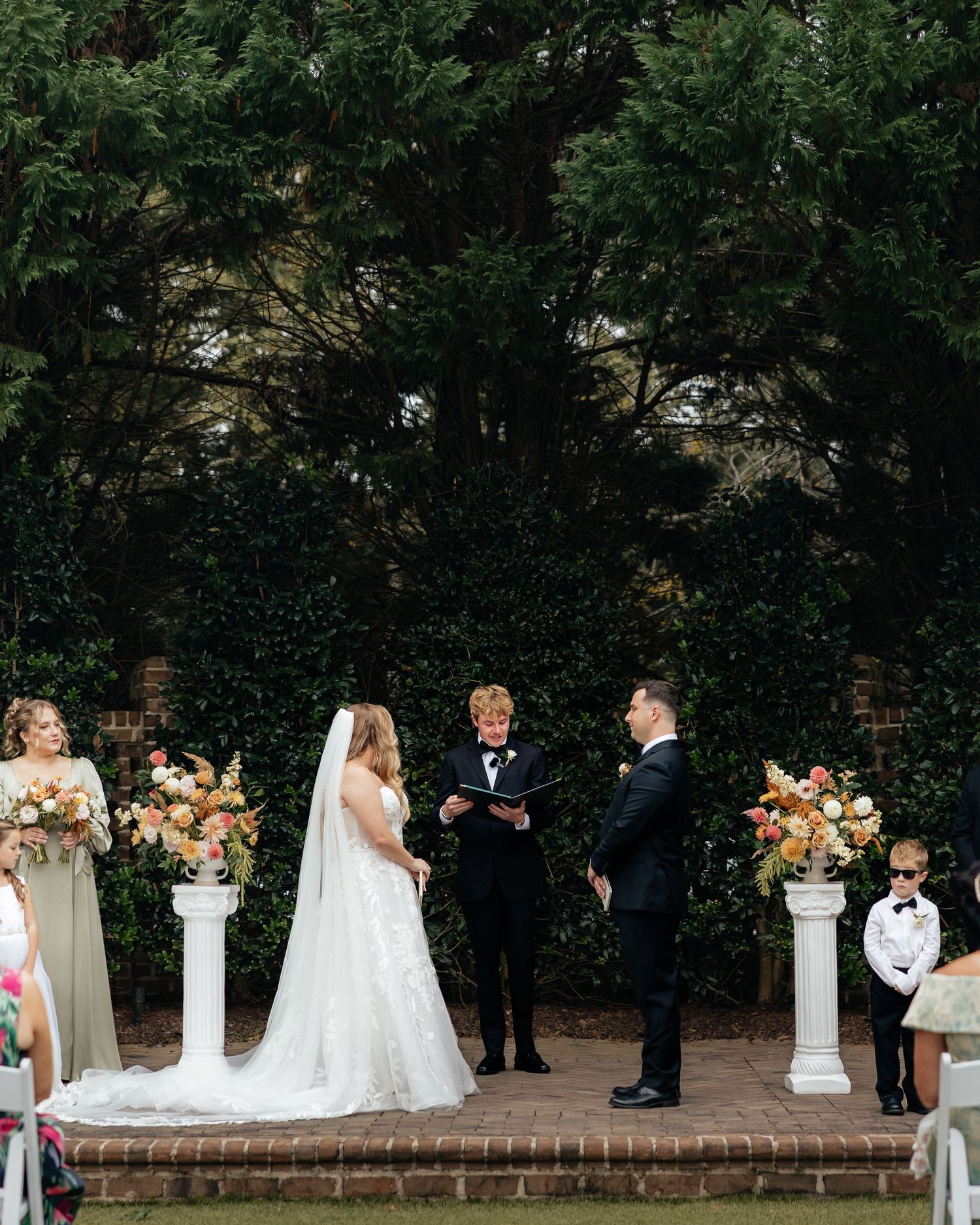 Wedding ceremony setup with white columns and floral arrangements along a brick wall in an outdoor garden setting.