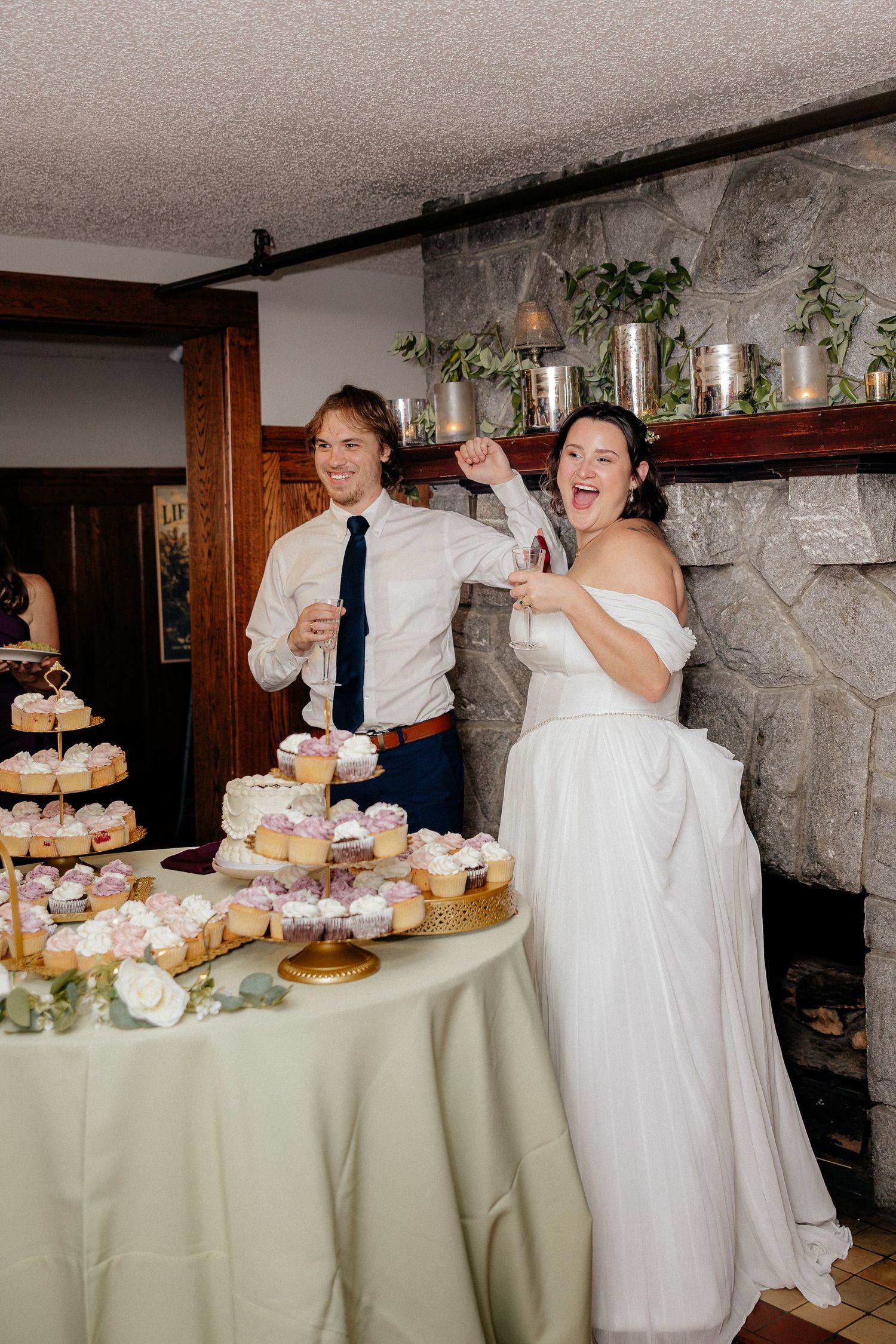 Laughing bride and groom cut their cupcake and cake display at a rustic stone-fireplace wedding reception.