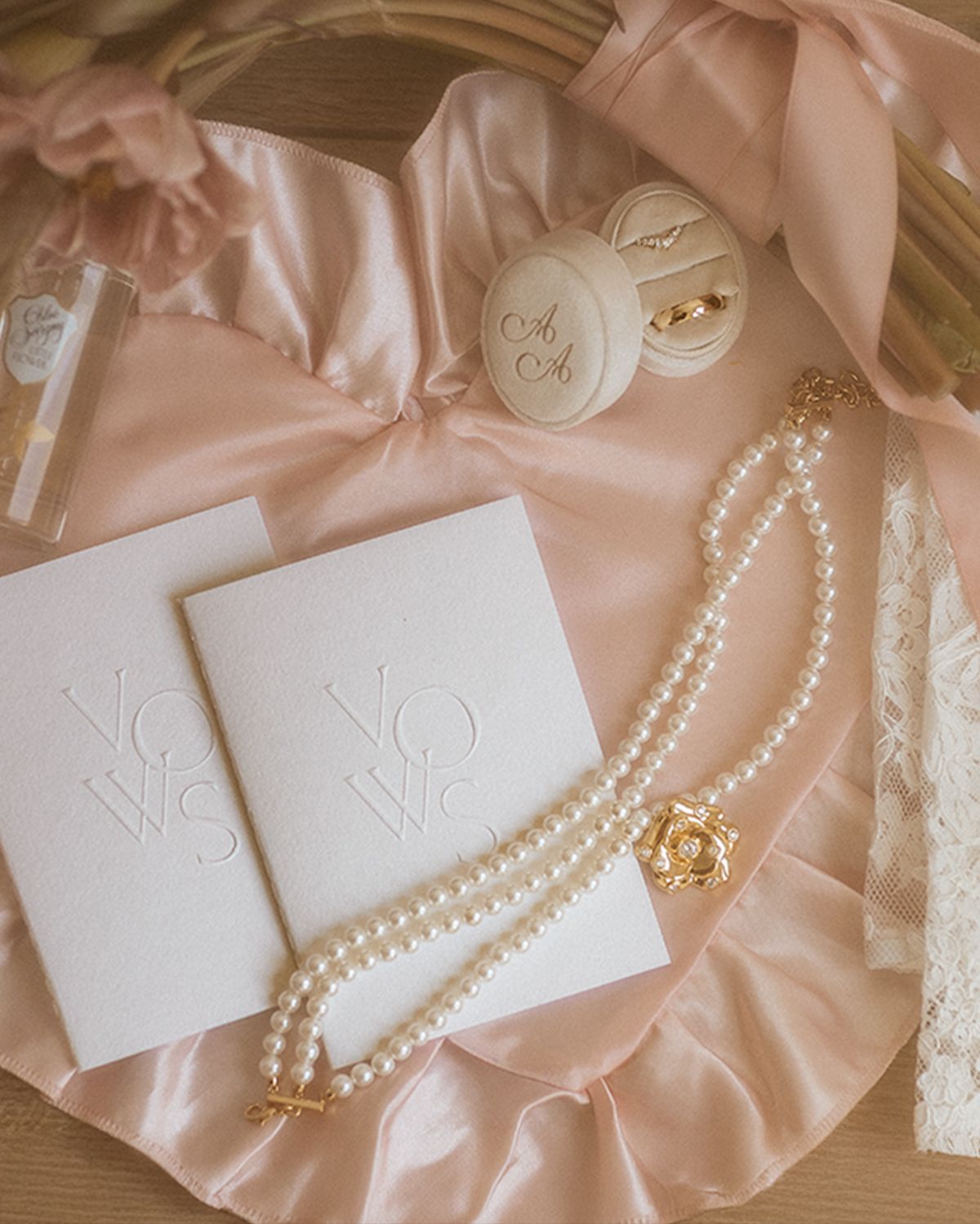 Elegant wedding flat lay with pearl necklace, vow books, ring box, and blush silk ribbon on pink satin fabric.